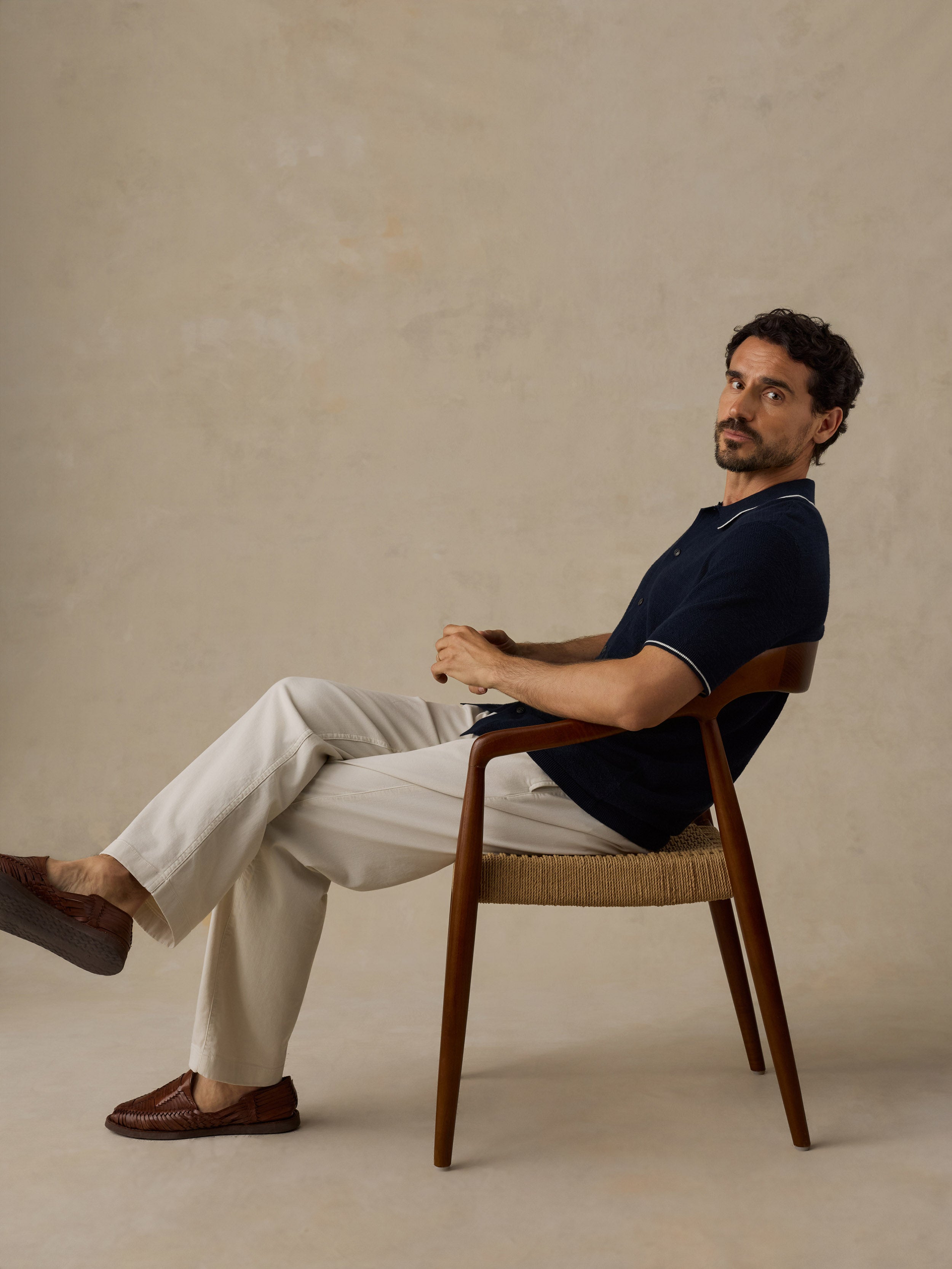 A man sits in a mid-century modern wooden armchair with a woven seat, wearing a navy blue short-sleeve polo shirt, cream trousers, and brown loafers against a textured beige studio backdrop.