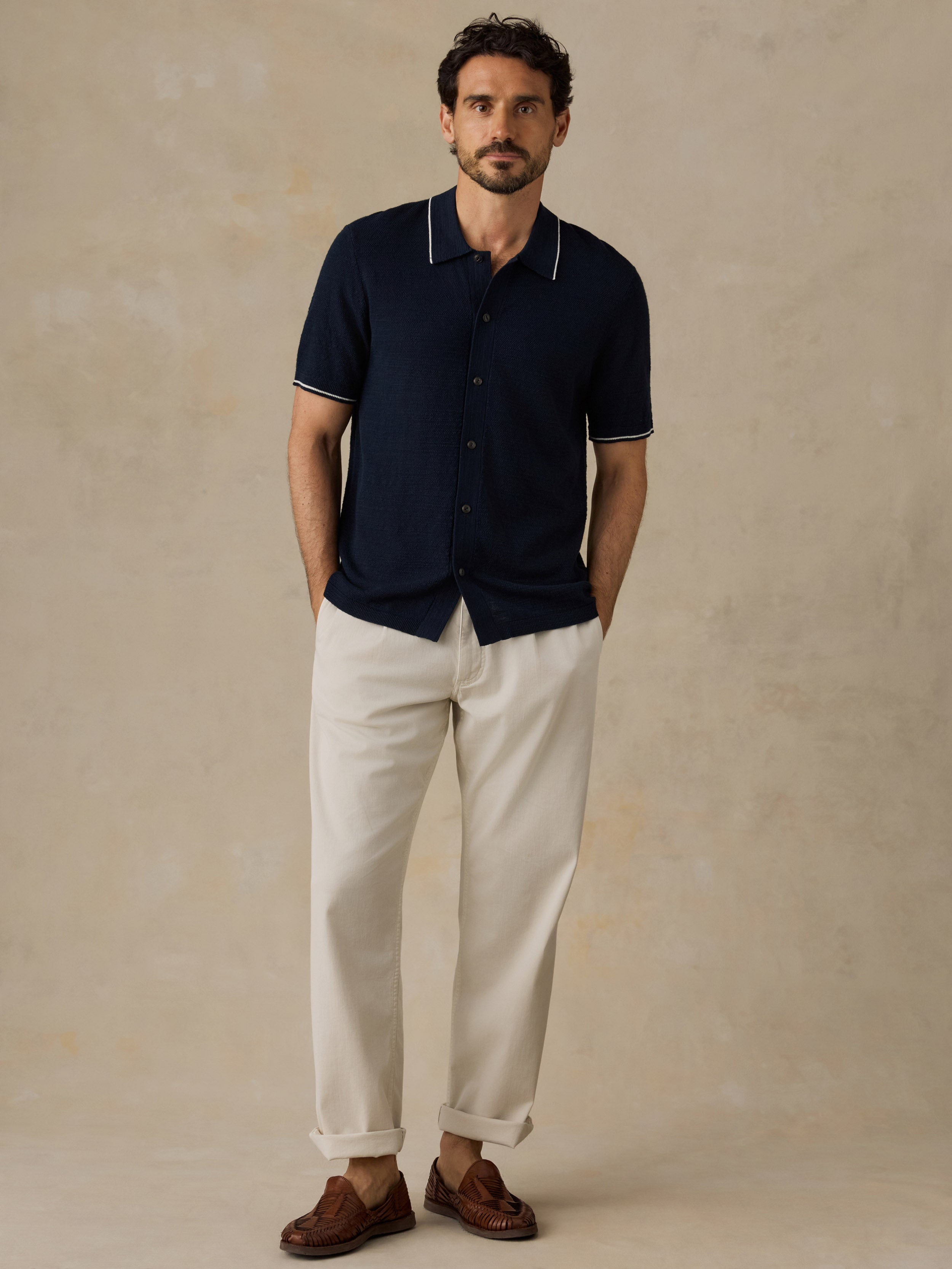 A man wearing a navy blue short-sleeve polo shirt with white piping, off-white trousers, and brown leather loafers, standing against a textured beige studio backdrop.