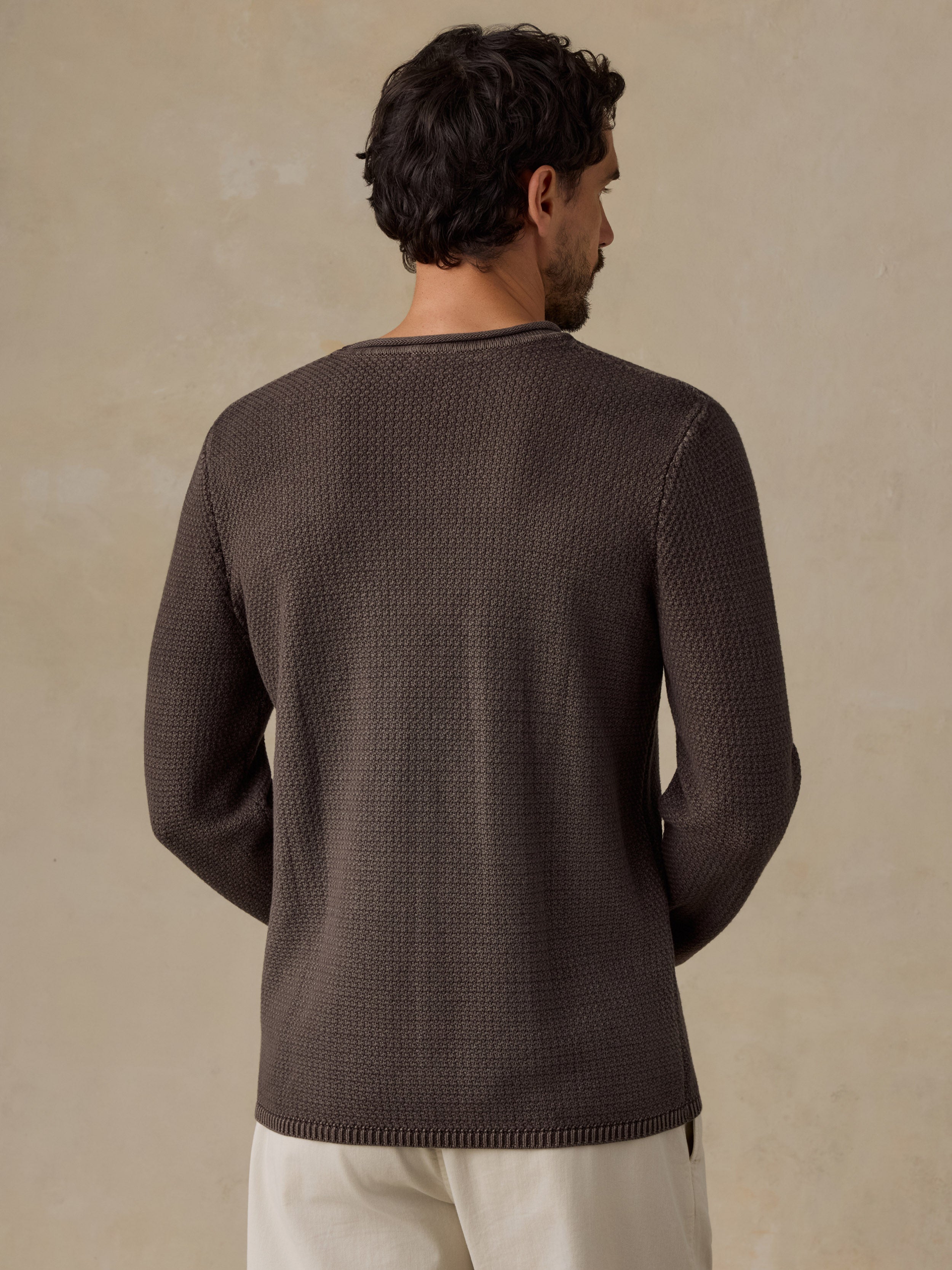 A man viewed from the back wearing a dark brown textured knit sweater and off-white trousers, standing against a warm beige background.