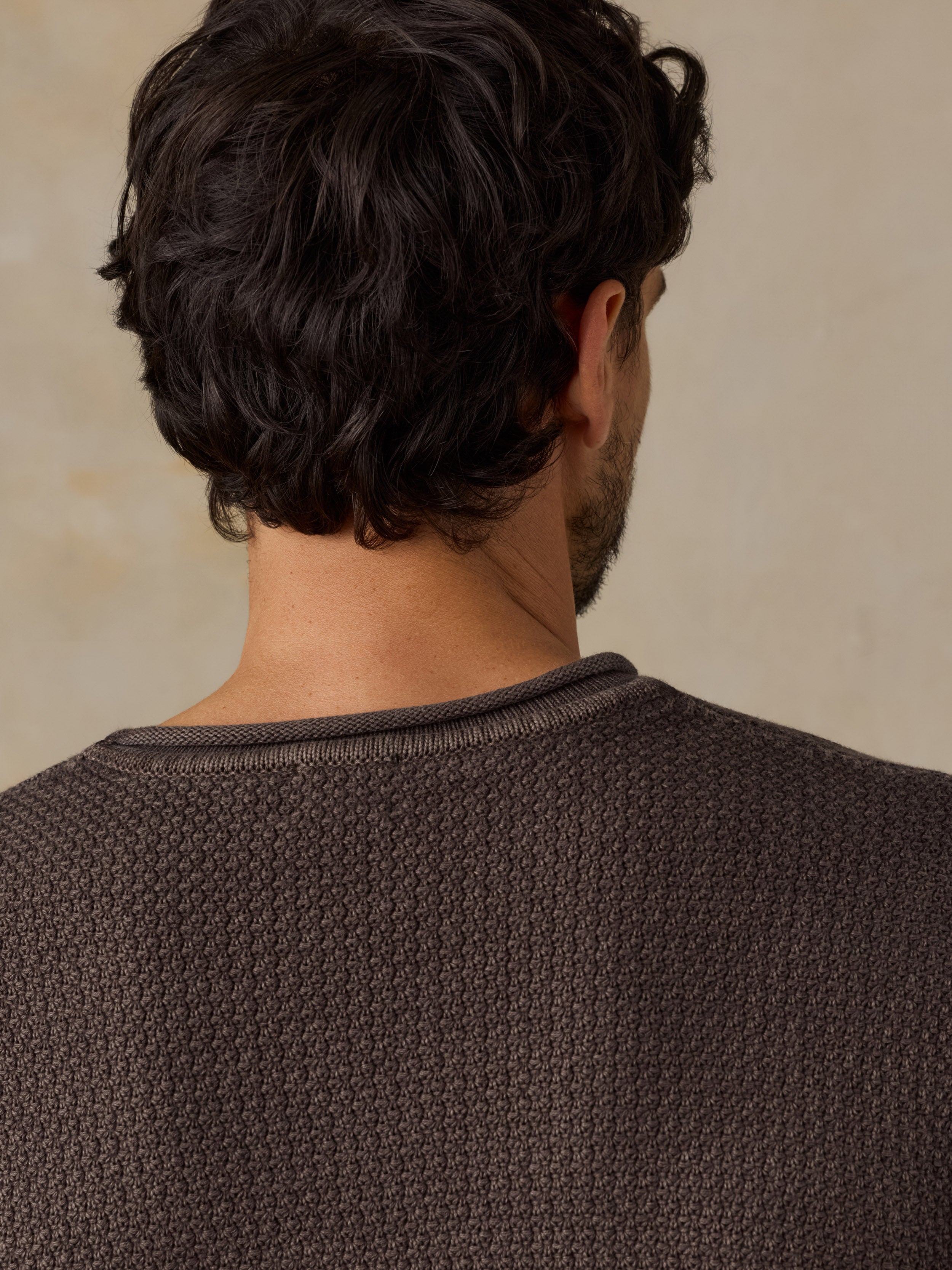 Close-up back view of a man wearing a dark brown textured knit crewneck sweater, showing the back of his head and neck against a warm beige background.