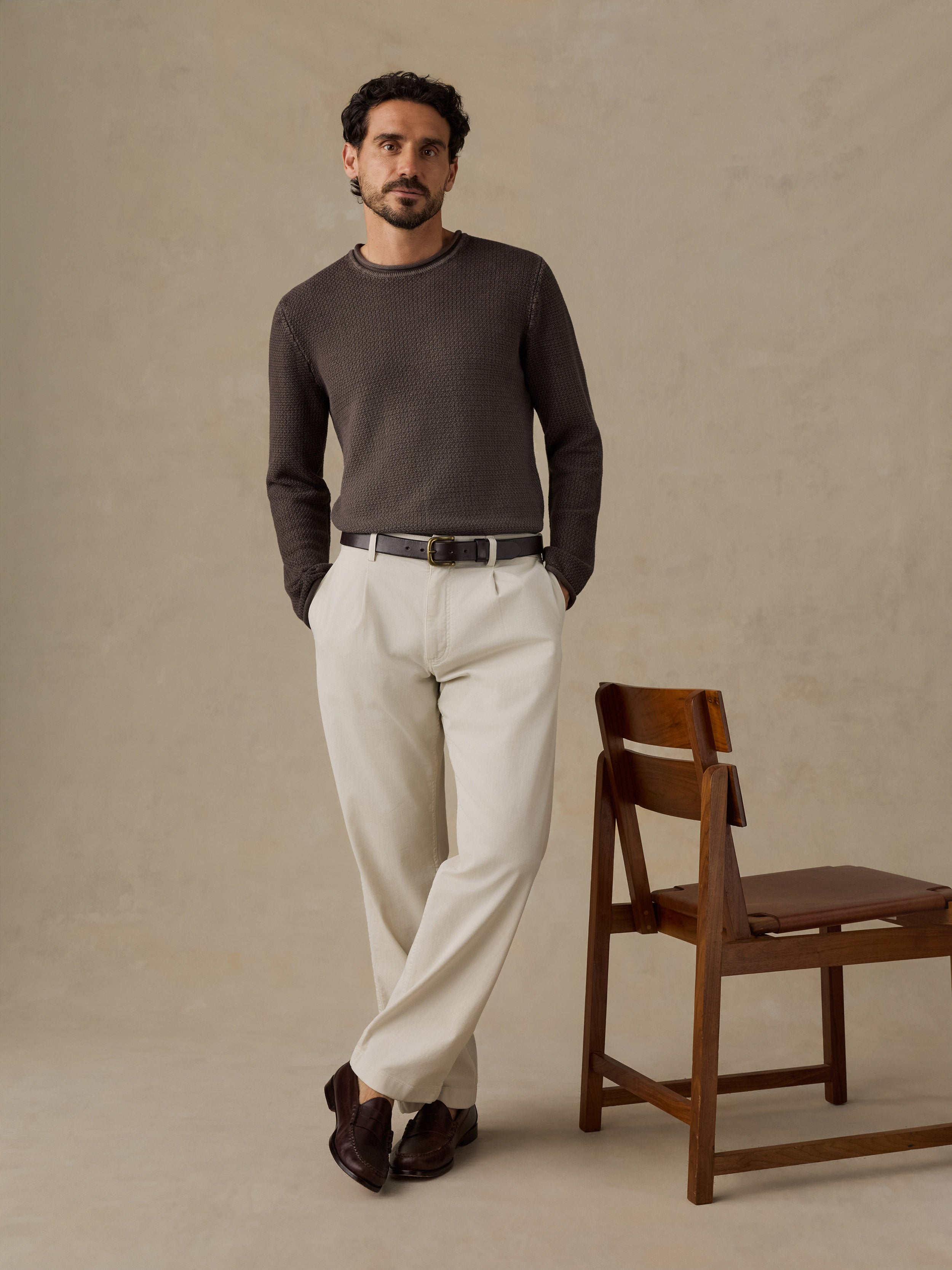 A man stands in a studio wearing a dark brown long-sleeve knit sweater, off-white trousers, a dark belt, and dark brown loafers, posed next to a wooden chair against a textured beige backdrop.