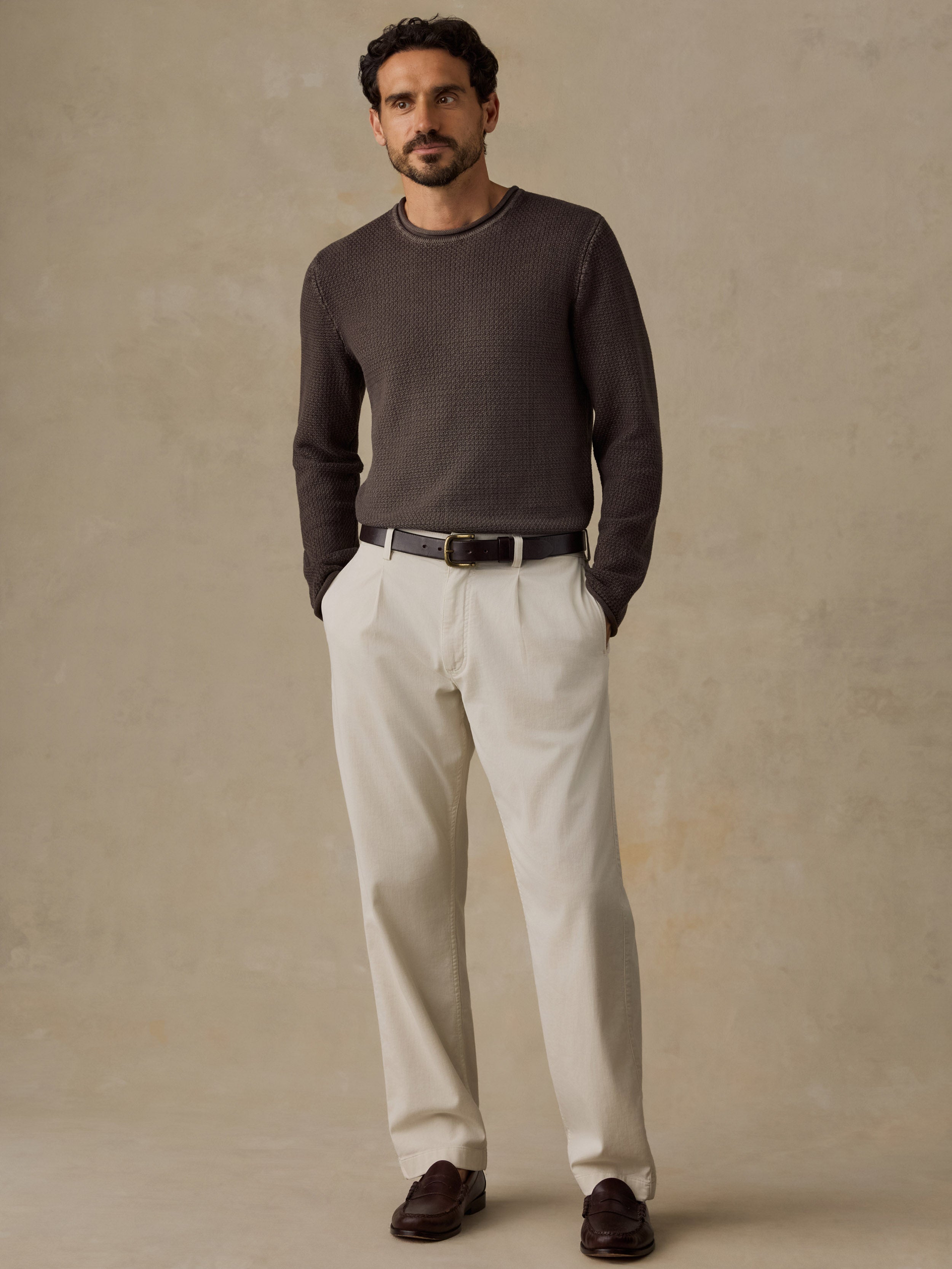 A man wearing a dark brown long-sleeve knit sweater, off-white trousers, a black belt, and dark brown loafers, standing against a textured warm beige studio backdrop.
