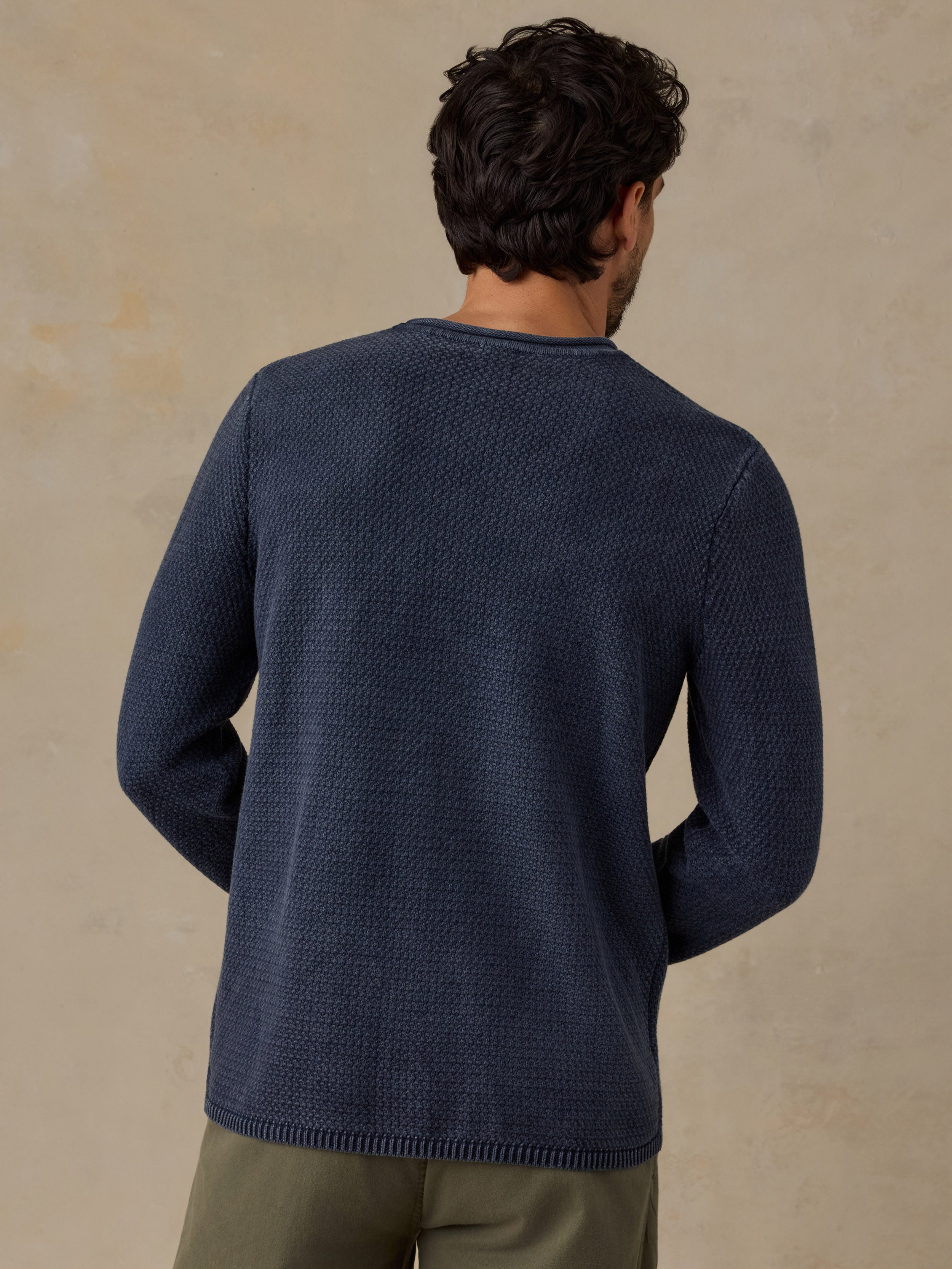 A man viewed from behind wearing a textured navy blue knit sweater and olive green pants, standing against a neutral beige background.