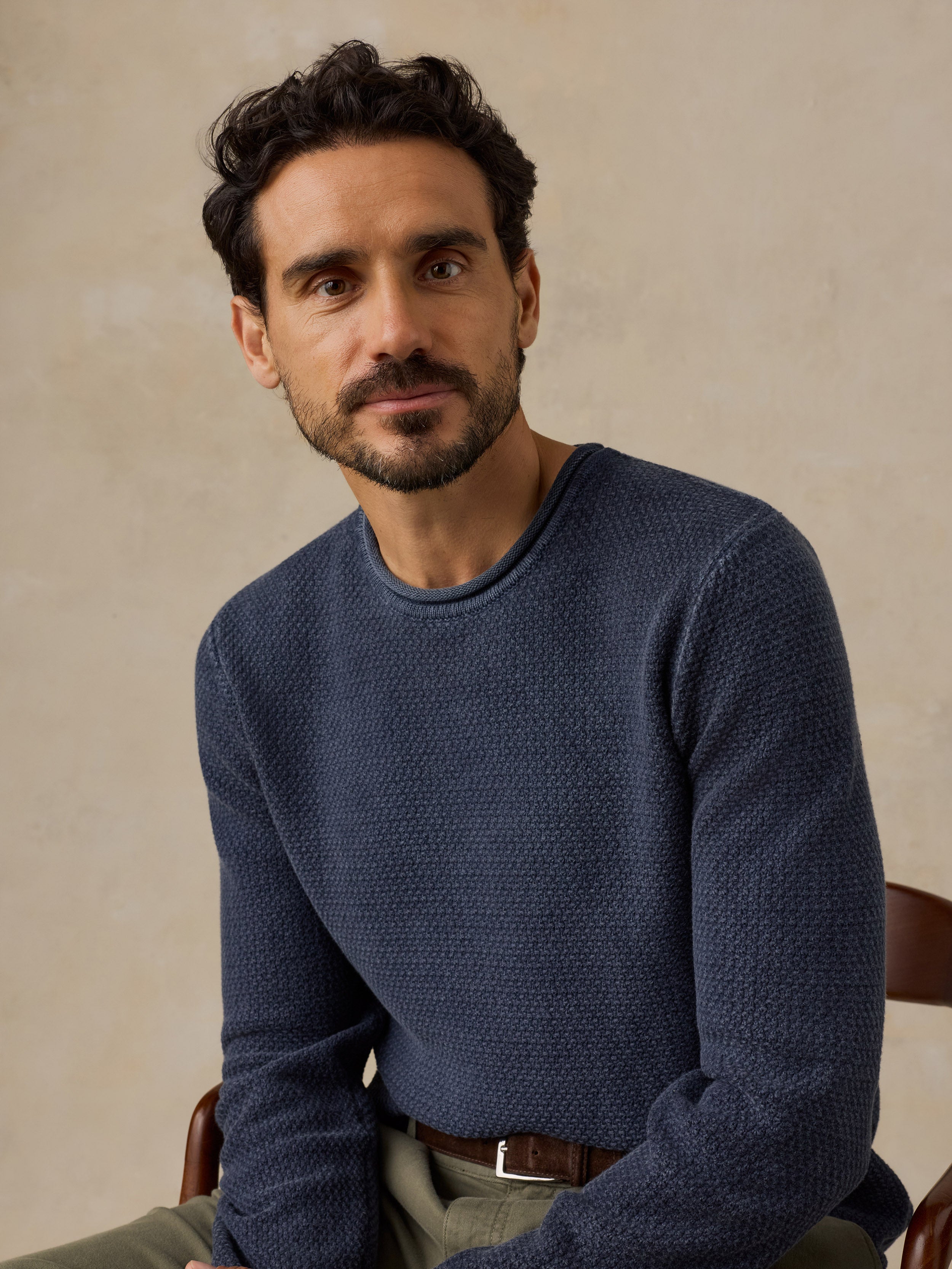 A man wearing a textured navy blue crewneck sweater, seated against a warm beige background.