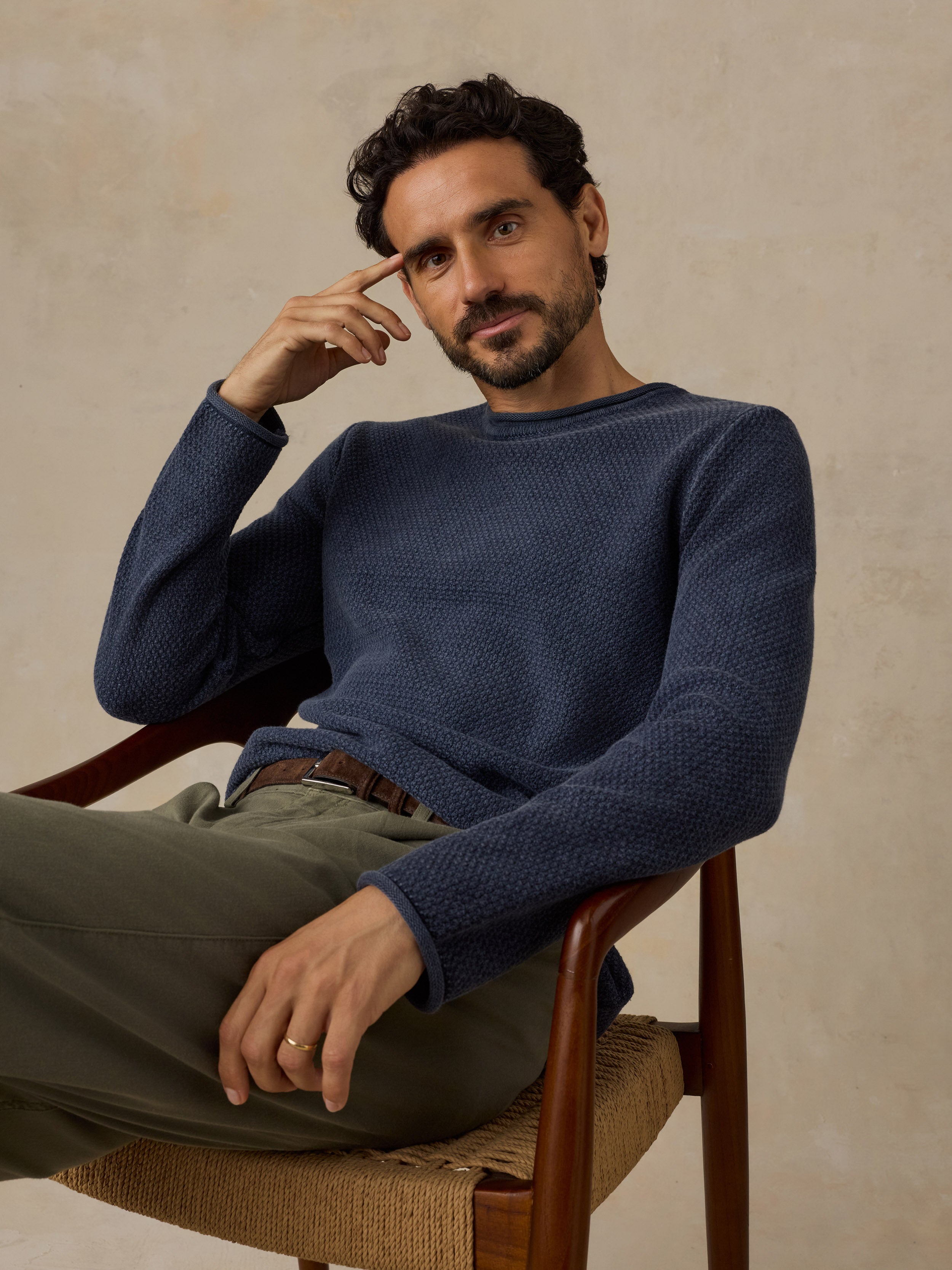 A man wearing a textured navy blue knit sweater and olive green trousers, sitting in a wooden chair against a warm beige background.