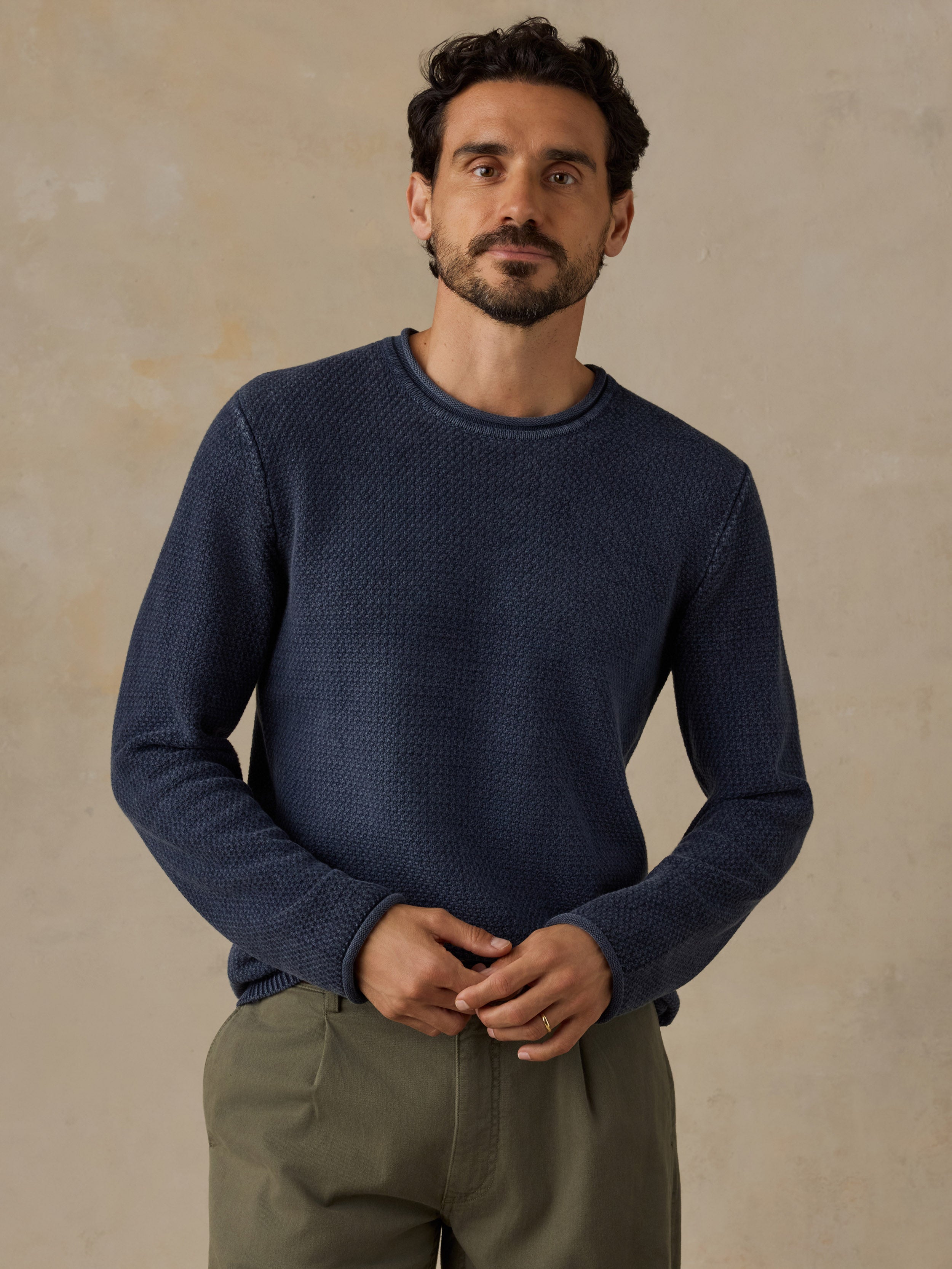 A man wearing a textured navy blue crewneck sweater and olive green trousers, standing against a neutral beige background.
