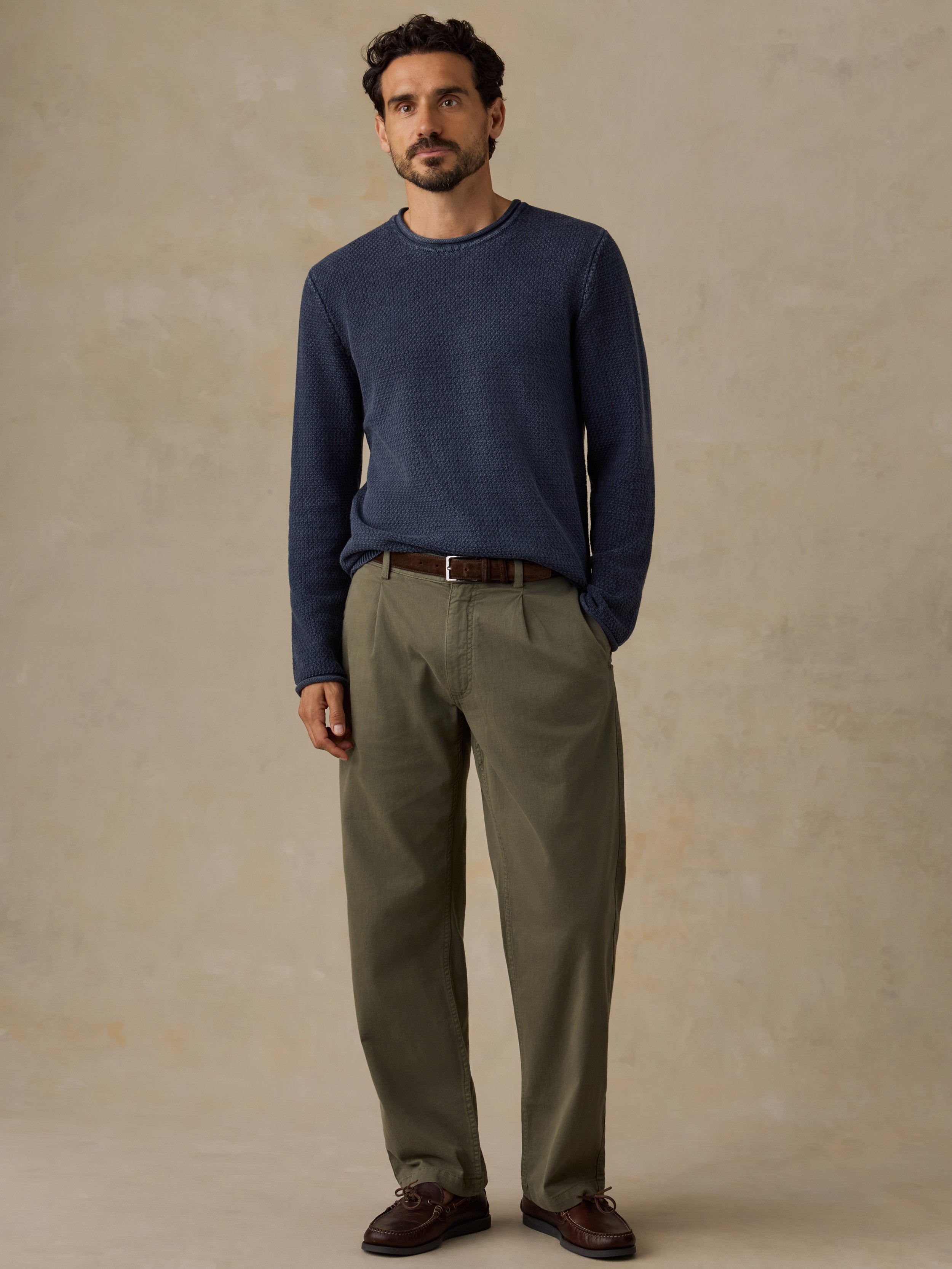 A man wearing a navy blue long-sleeve knit sweater, olive green trousers, a brown belt, and dark brown shoes, standing against a textured beige studio background.