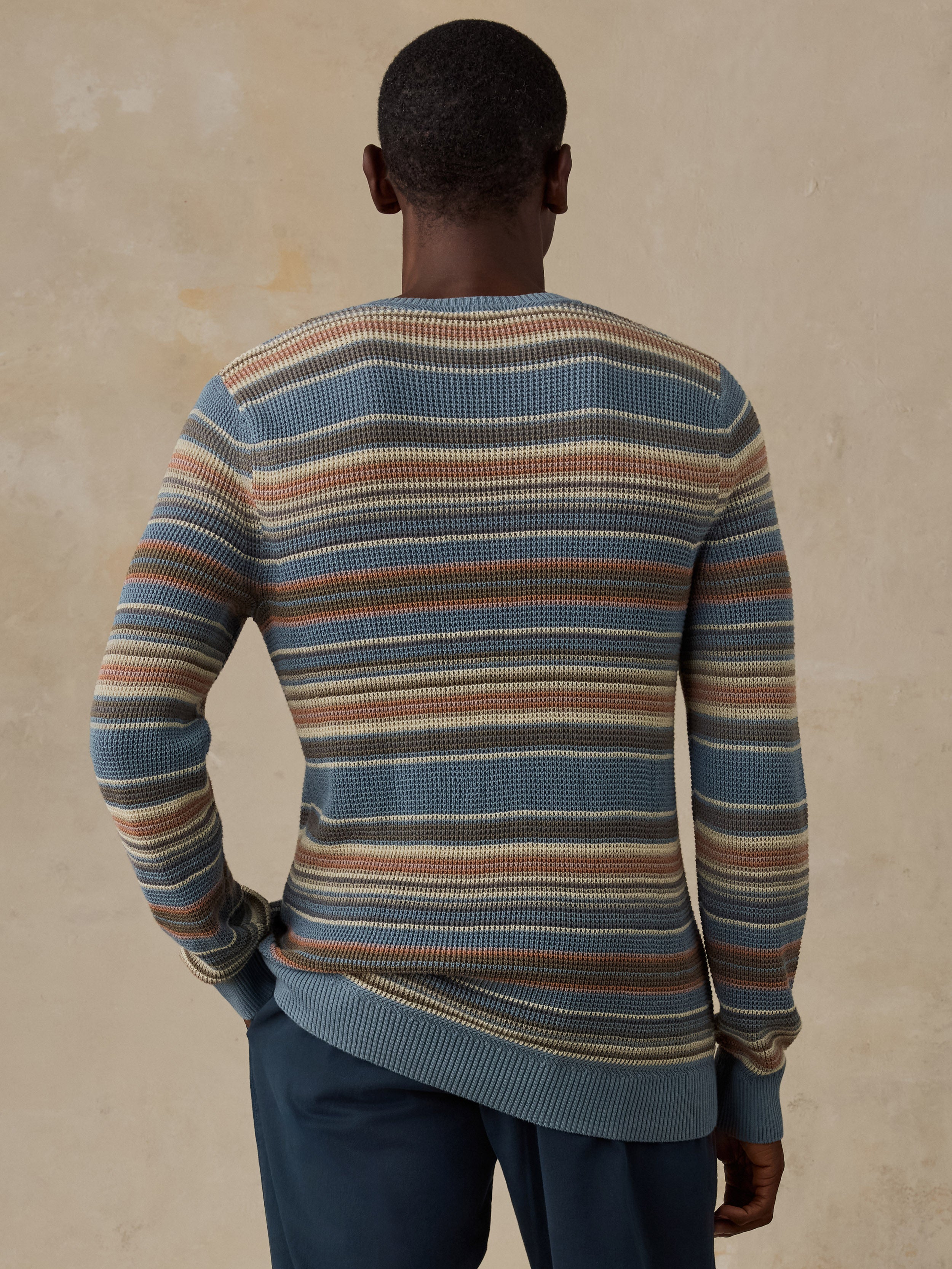 A man wearing a blue, beige, and brown horizontally striped crewneck sweater and dark navy trousers, standing against a beige background.