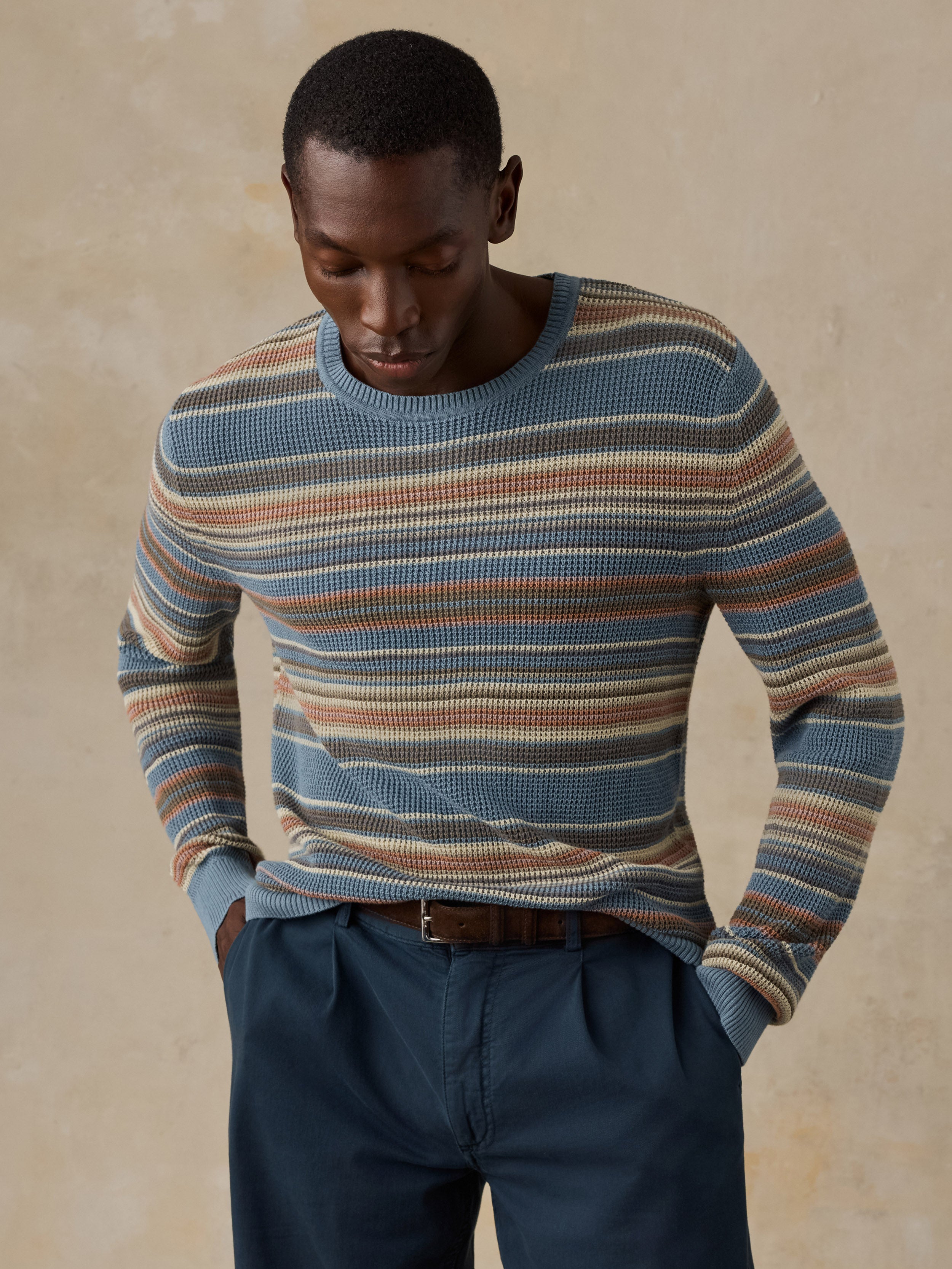 A man wearing a blue, beige, and brown horizontally striped crewneck sweater and dark navy trousers, standing against a beige background.