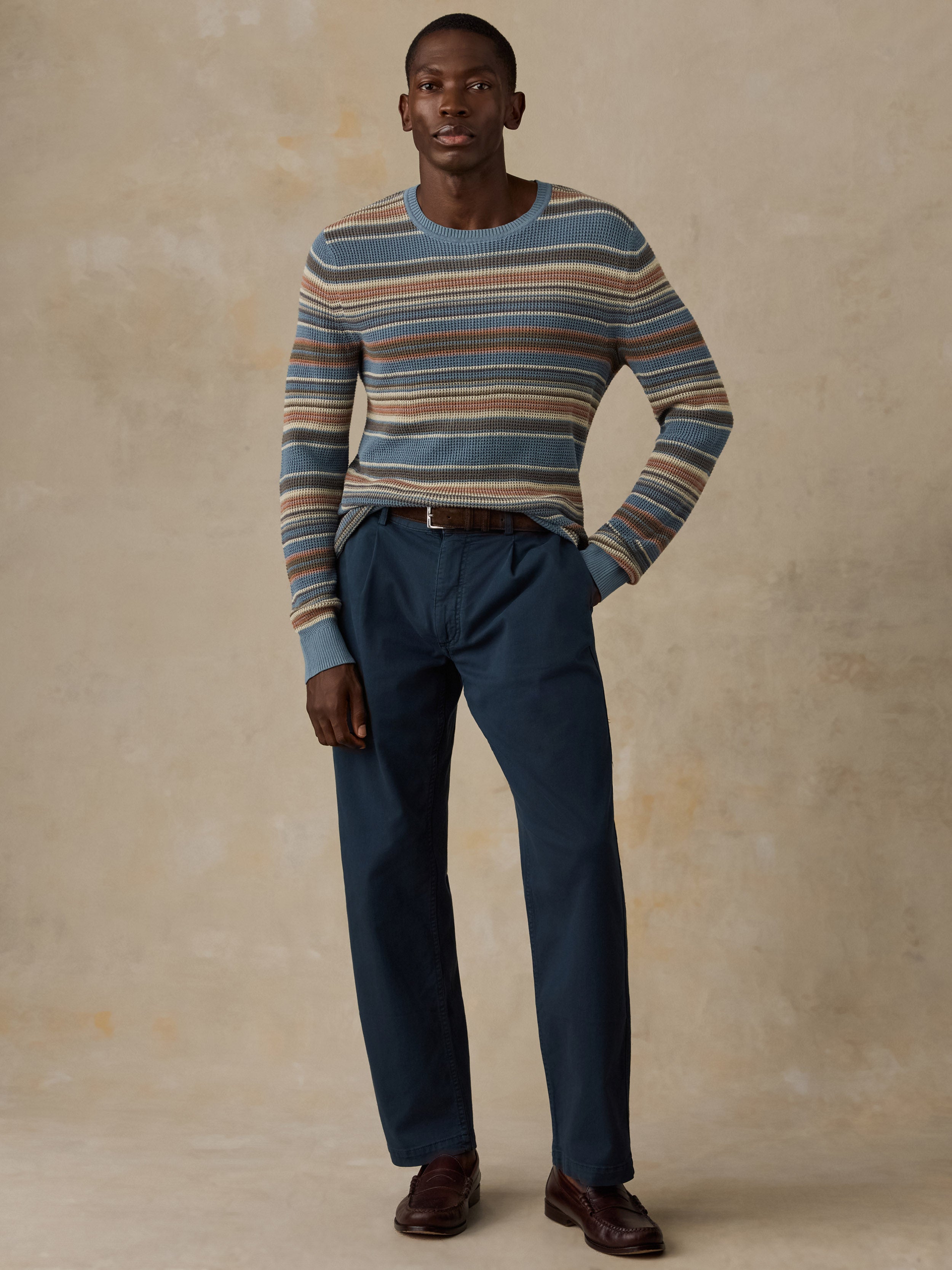 A man wearing a blue, beige, and brown horizontally striped crewneck sweater and dark navy trousers, standing against a beige background.