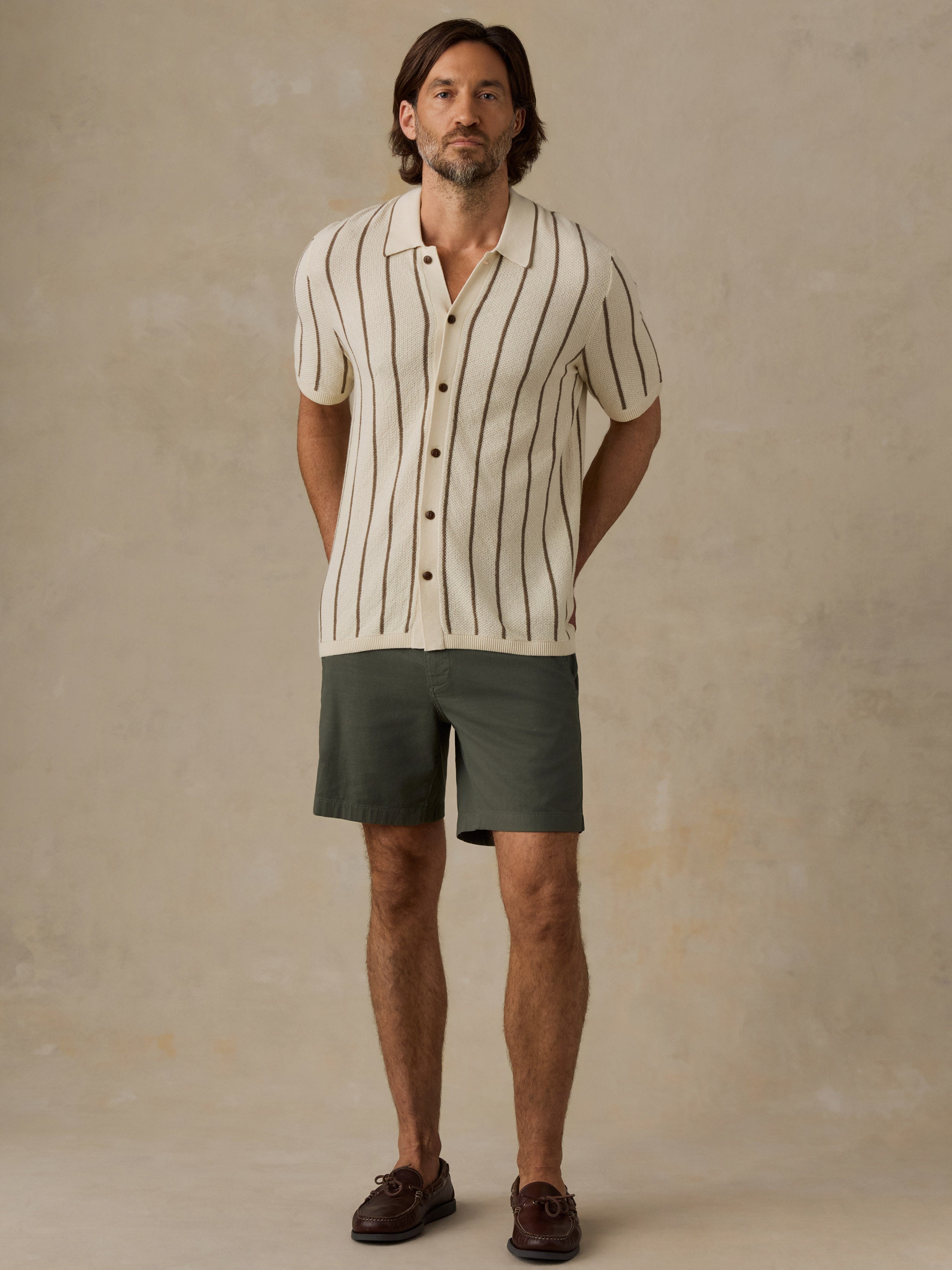 A man modeling a short-sleeve button-up shirt with thin vertical black stripes on an off-white base, paired with dark olive green shorts and brown leather shoes, against a textured beige studio backdrop.