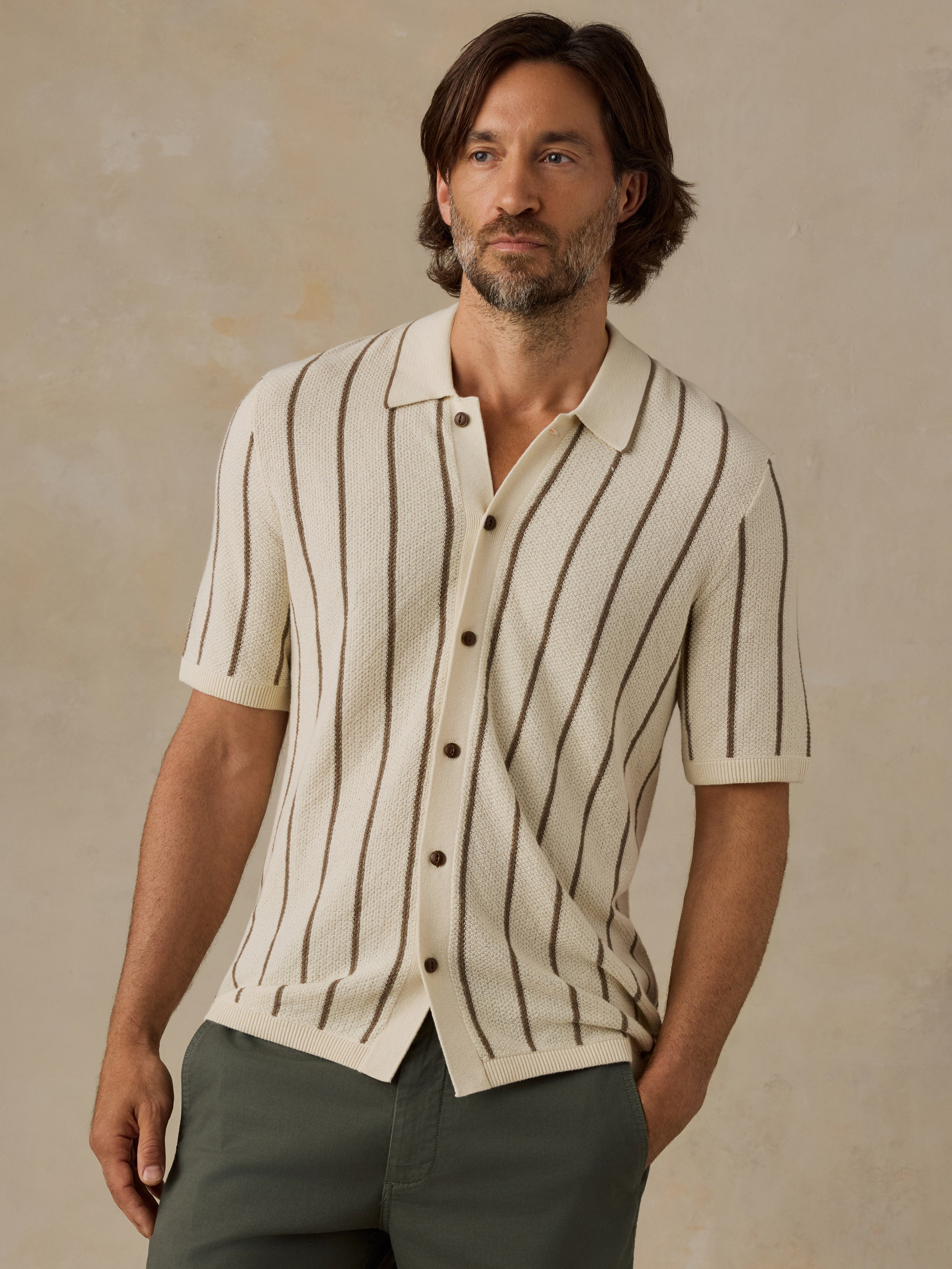 A man wearing a cream-colored short-sleeve knit button-up shirt with vertical brown stripes, paired with brown trousers, standing against a neutral beige background.