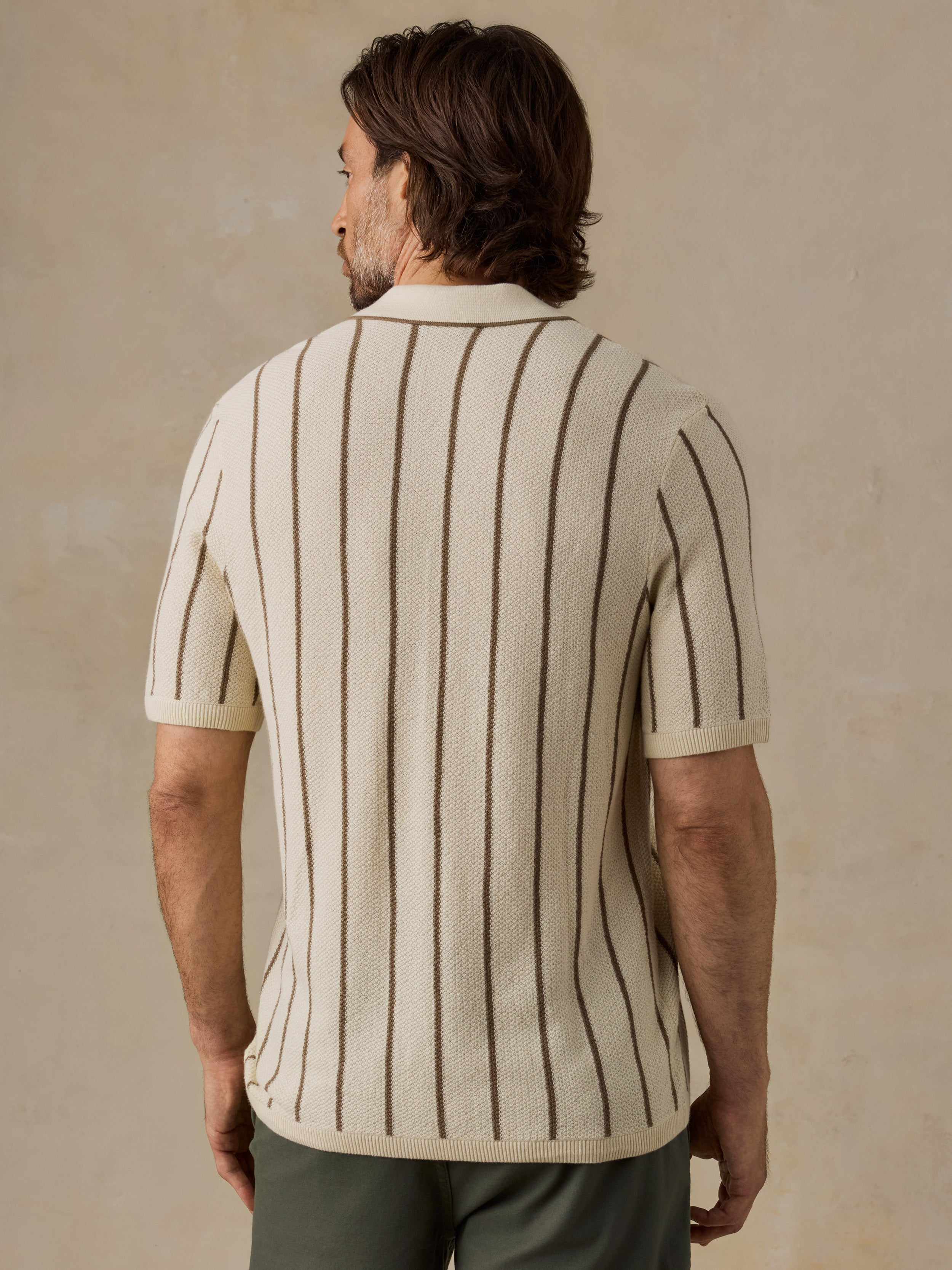 A man viewed from the back wearing a cream knit short-sleeve button-up shirt with vertical brown stripes, paired with brown trousers, against a textured beige background.