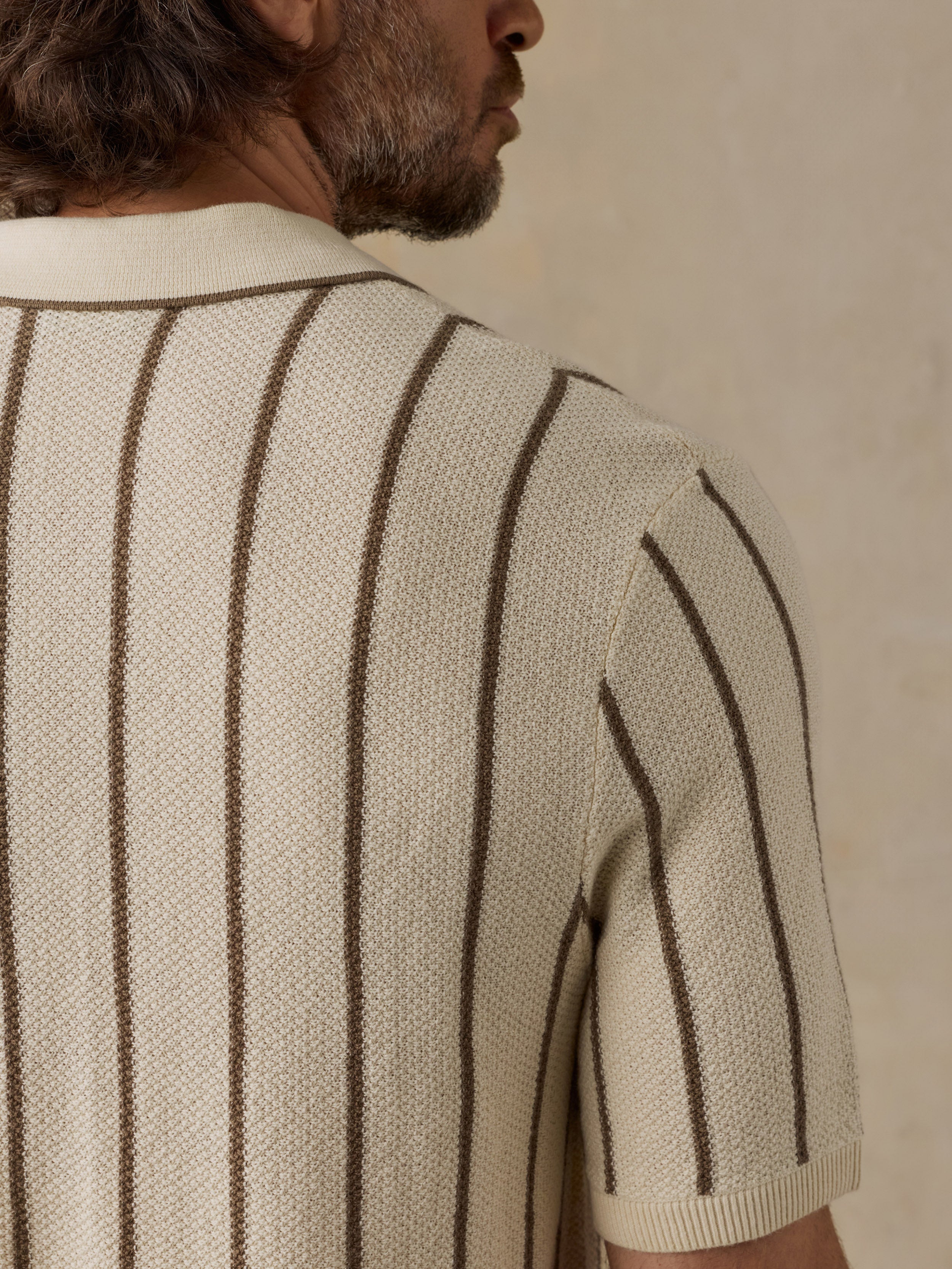Close-up back view of a man wearing a cream knit short-sleeve sweater with dark brown vertical pinstripes, set against a neutral beige background.