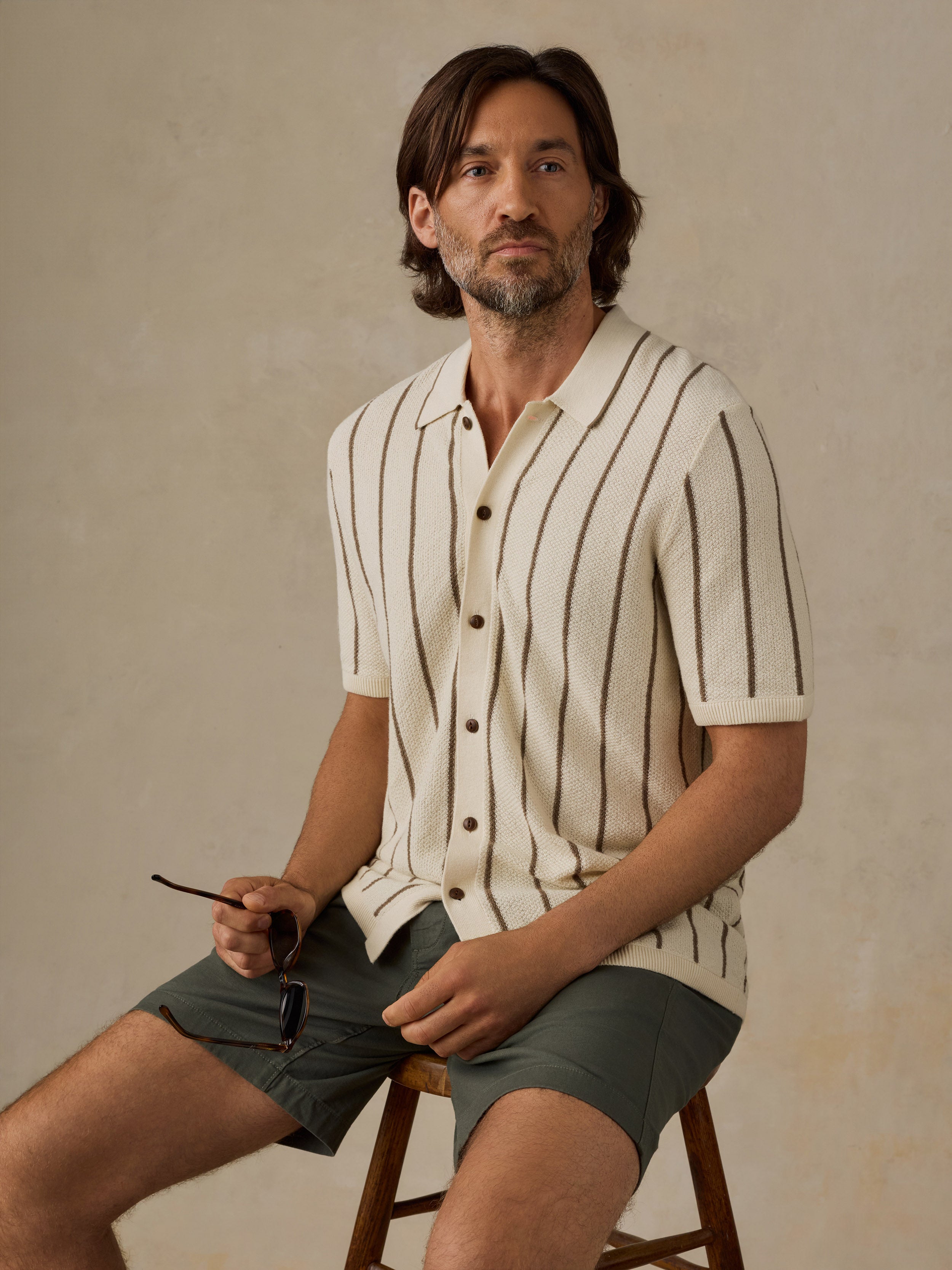 A man wearing a cream short-sleeve knit button-up shirt with thin dark vertical stripes and olive green shorts, sitting on a wooden stool against a textured beige studio background.