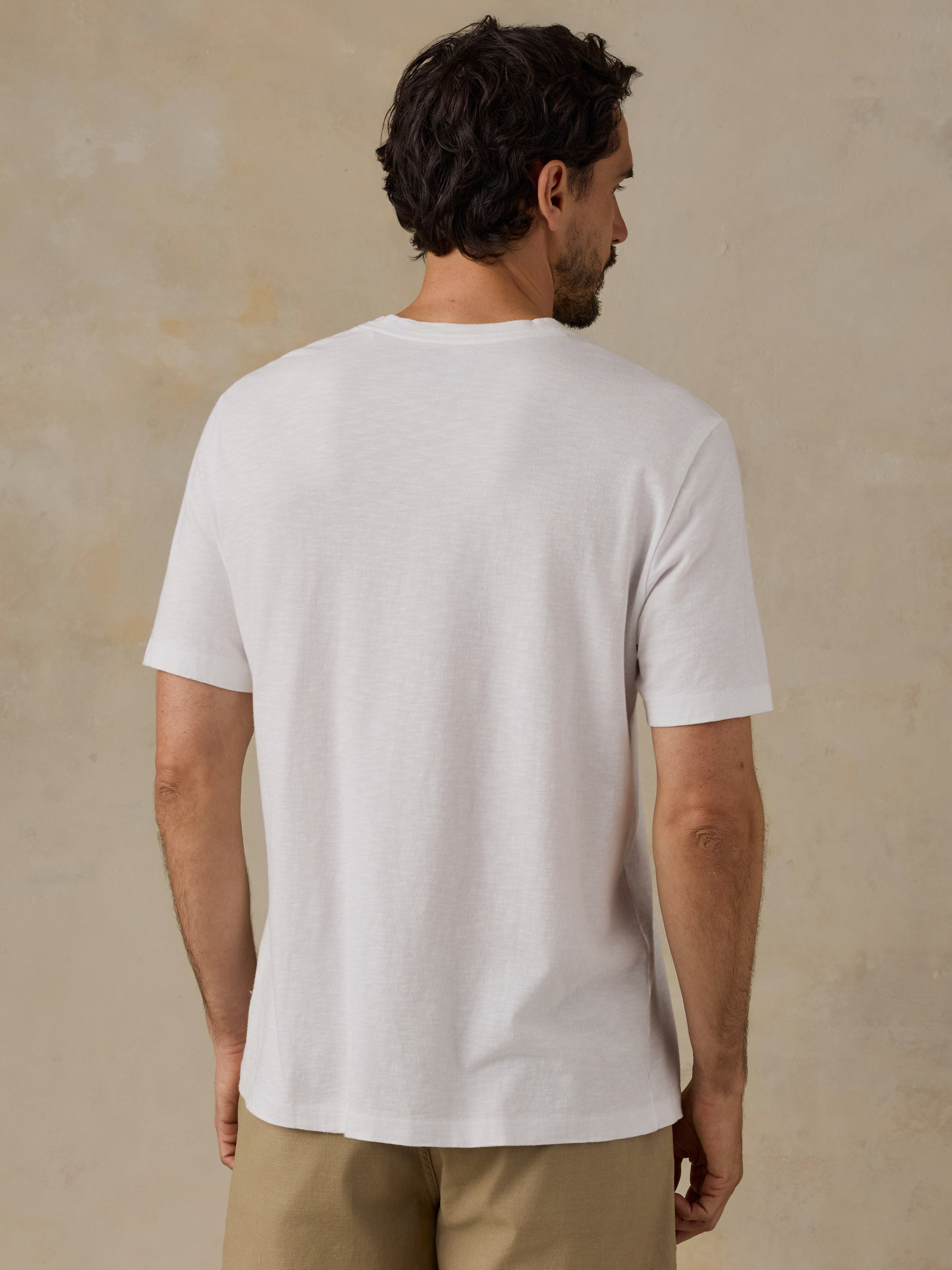 A man viewed from behind wearing a plain white short-sleeve crew-neck t-shirt and khaki pants, standing against a textured beige wall.