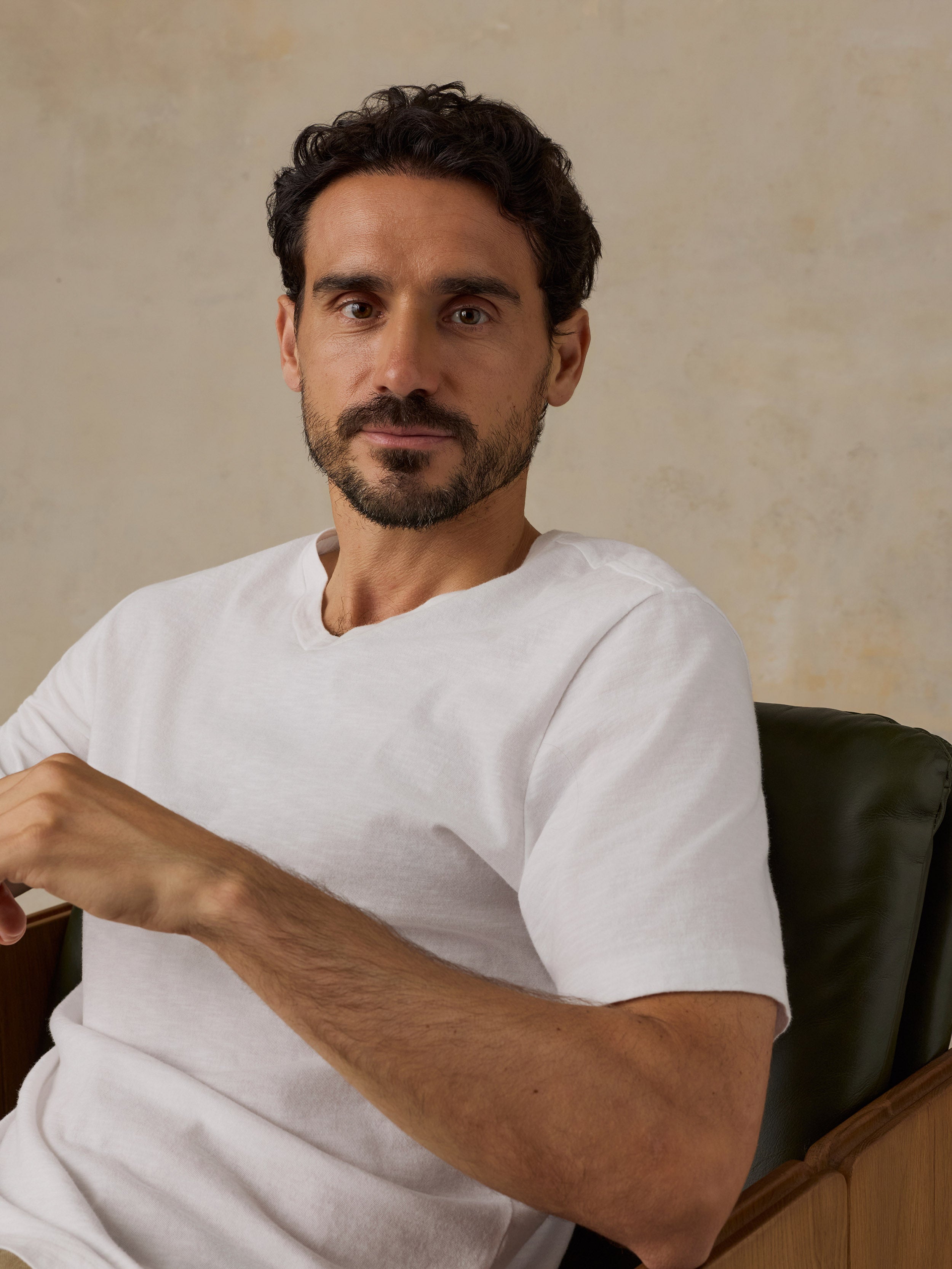 A man with dark curly hair and a beard wearing a white short-sleeve crewneck t-shirt, sitting in a dark green leather chair against a textured beige wall.