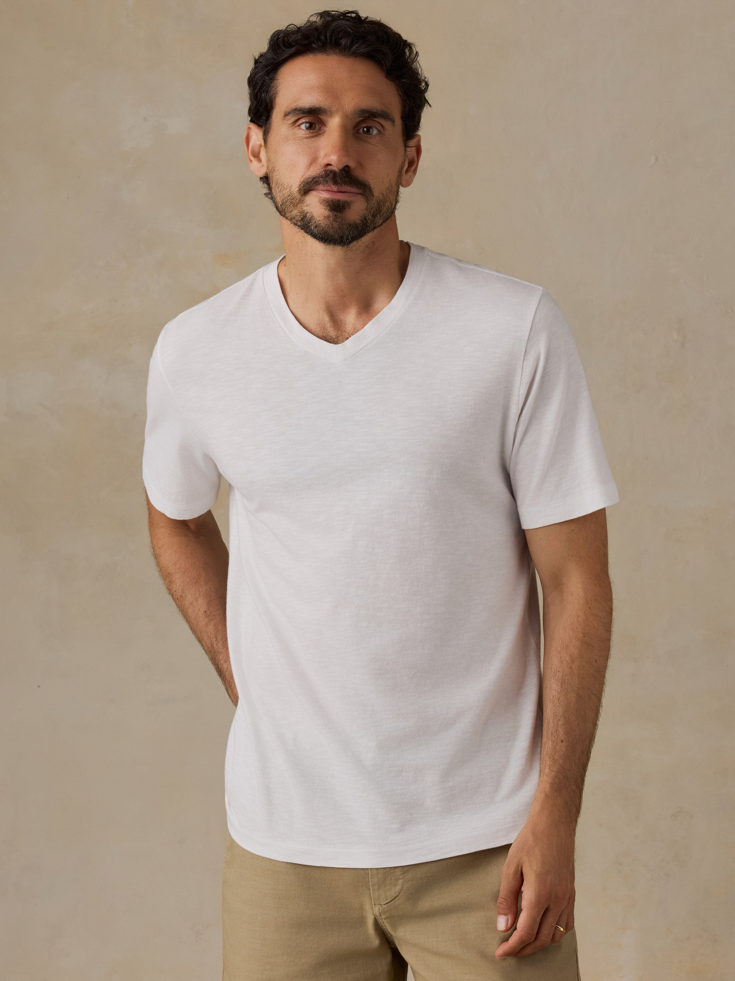 A man wearing a white short-sleeve v-neck t-shirt and khaki pants, standing against a textured beige wall.