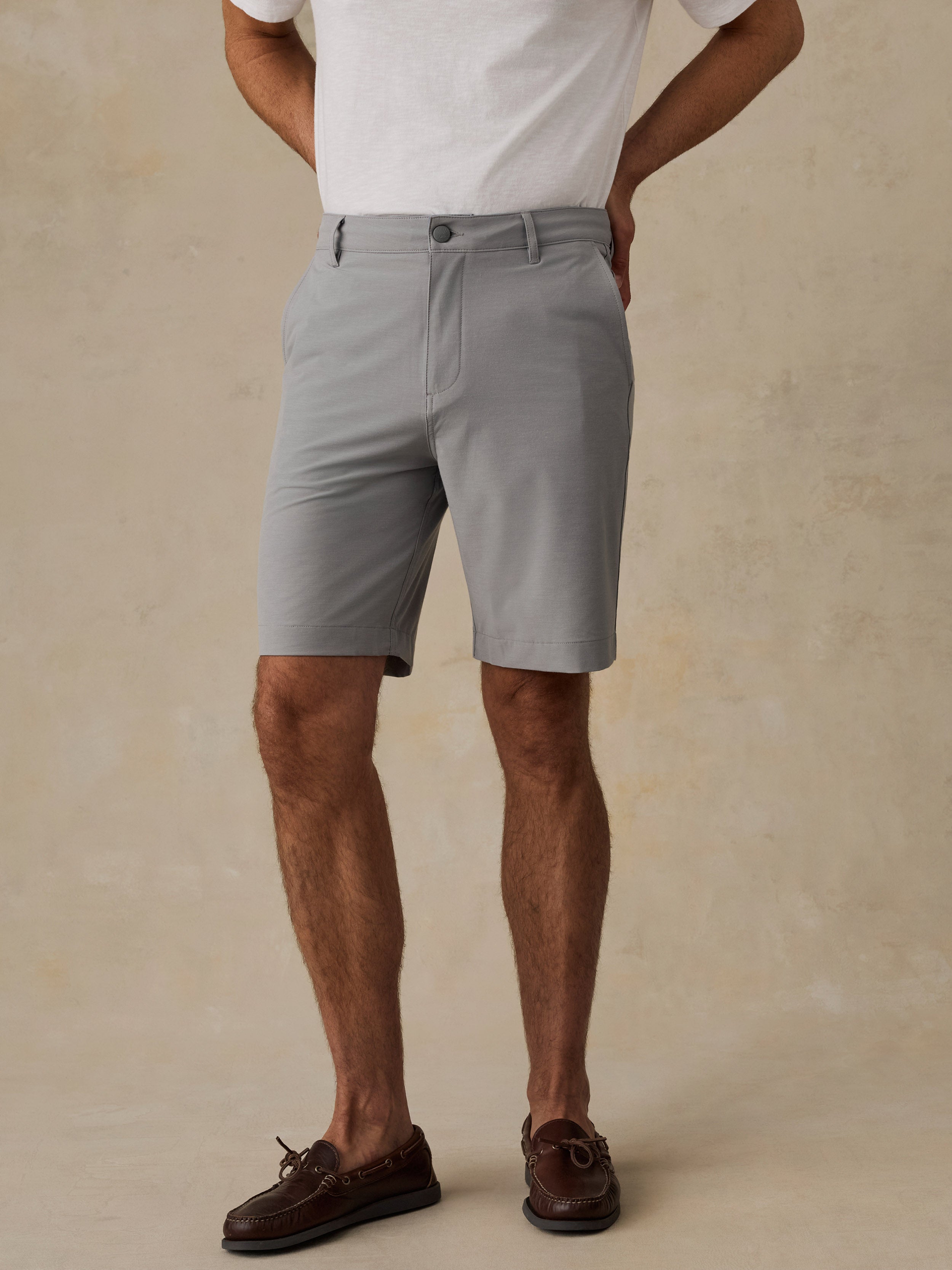 A man wearing light gray chino shorts, a white t-shirt, and brown boat shoes, standing against a textured beige background.