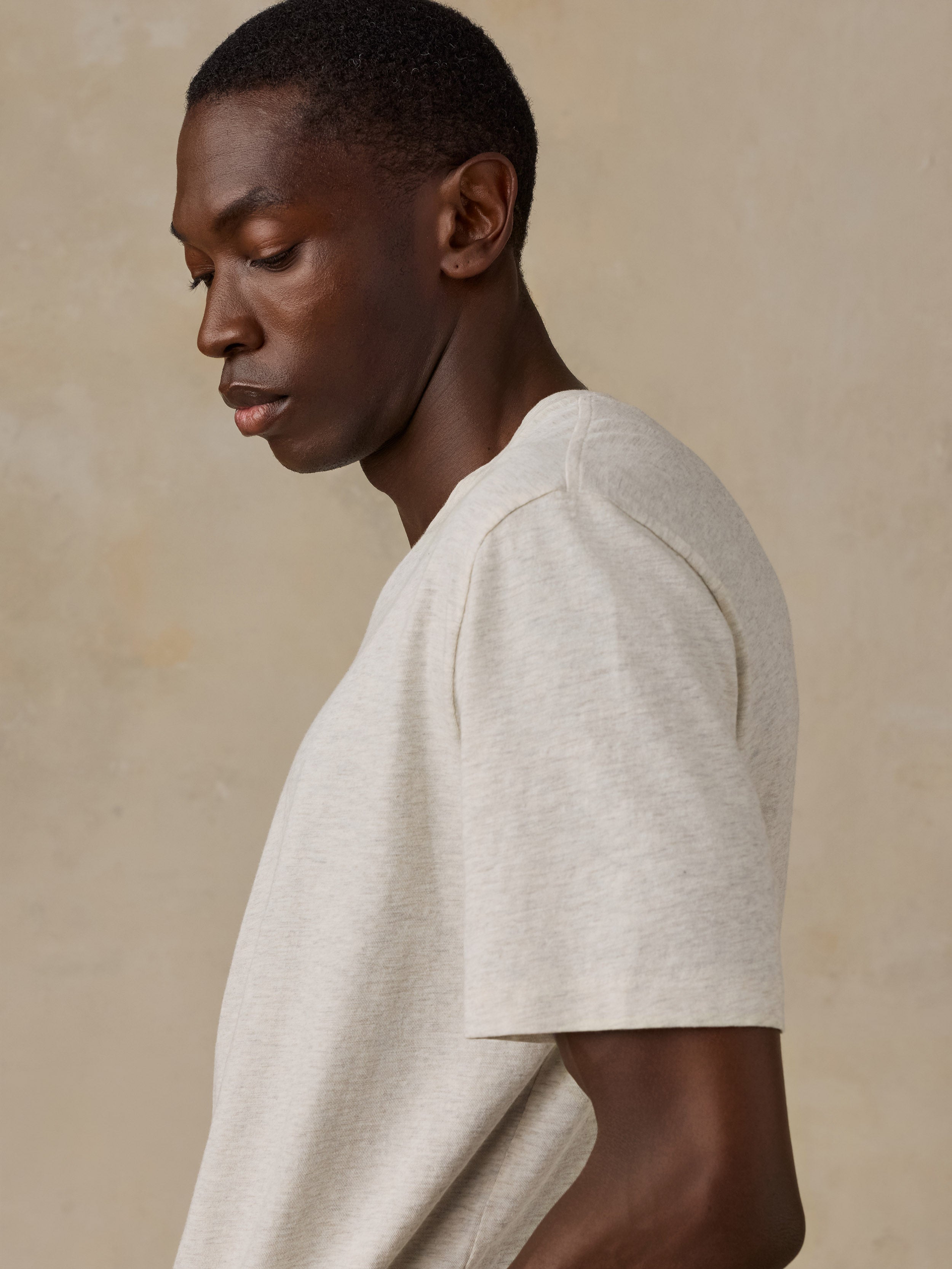 A man in profile wearing a short-sleeve crew-neck t-shirt in a light oatmeal beige color, set against a textured beige background.
