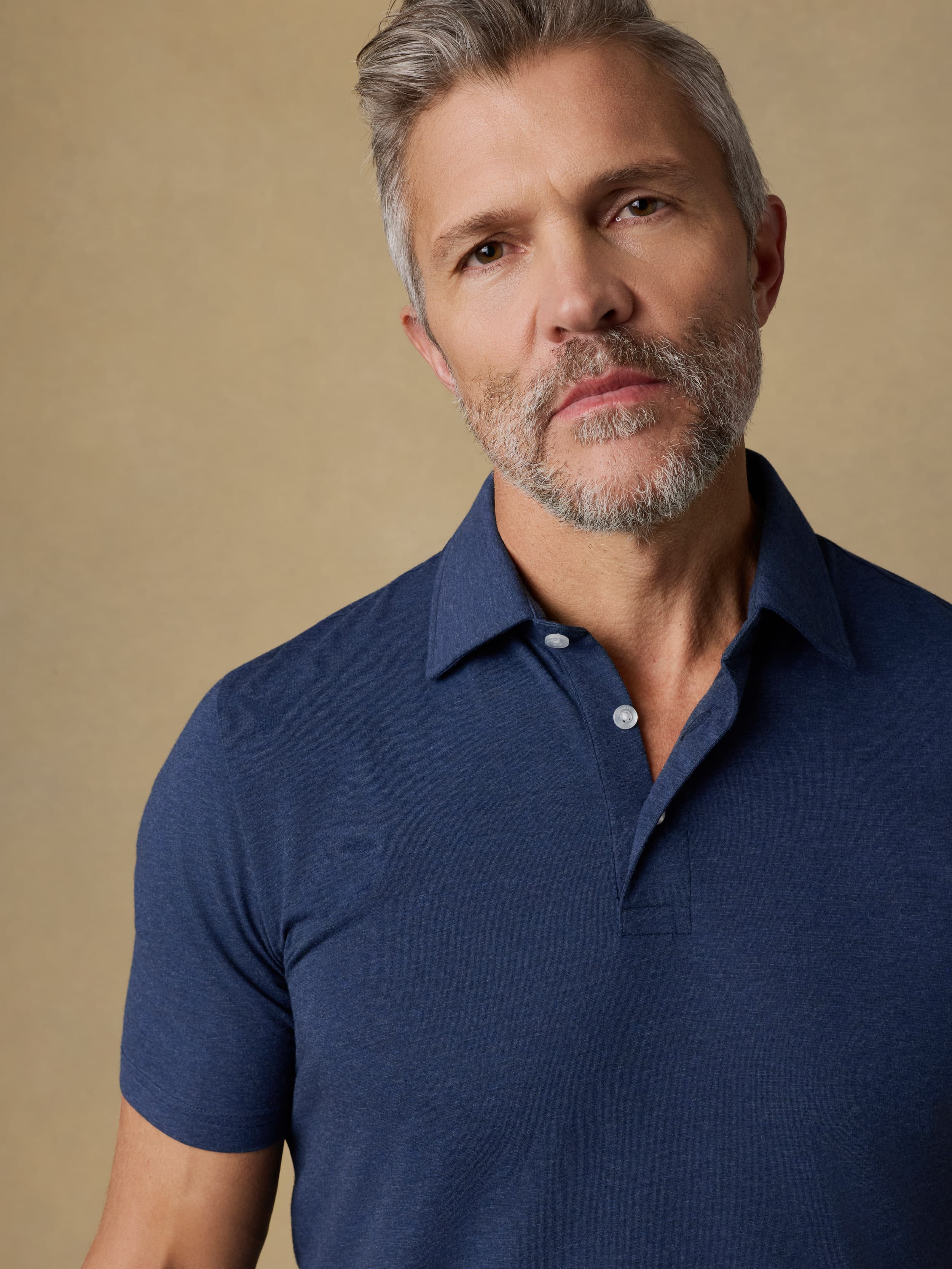 A close-up shot of a man wearing a short-sleeve polo shirt in heathered navy blue, set against a solid warm beige background.
