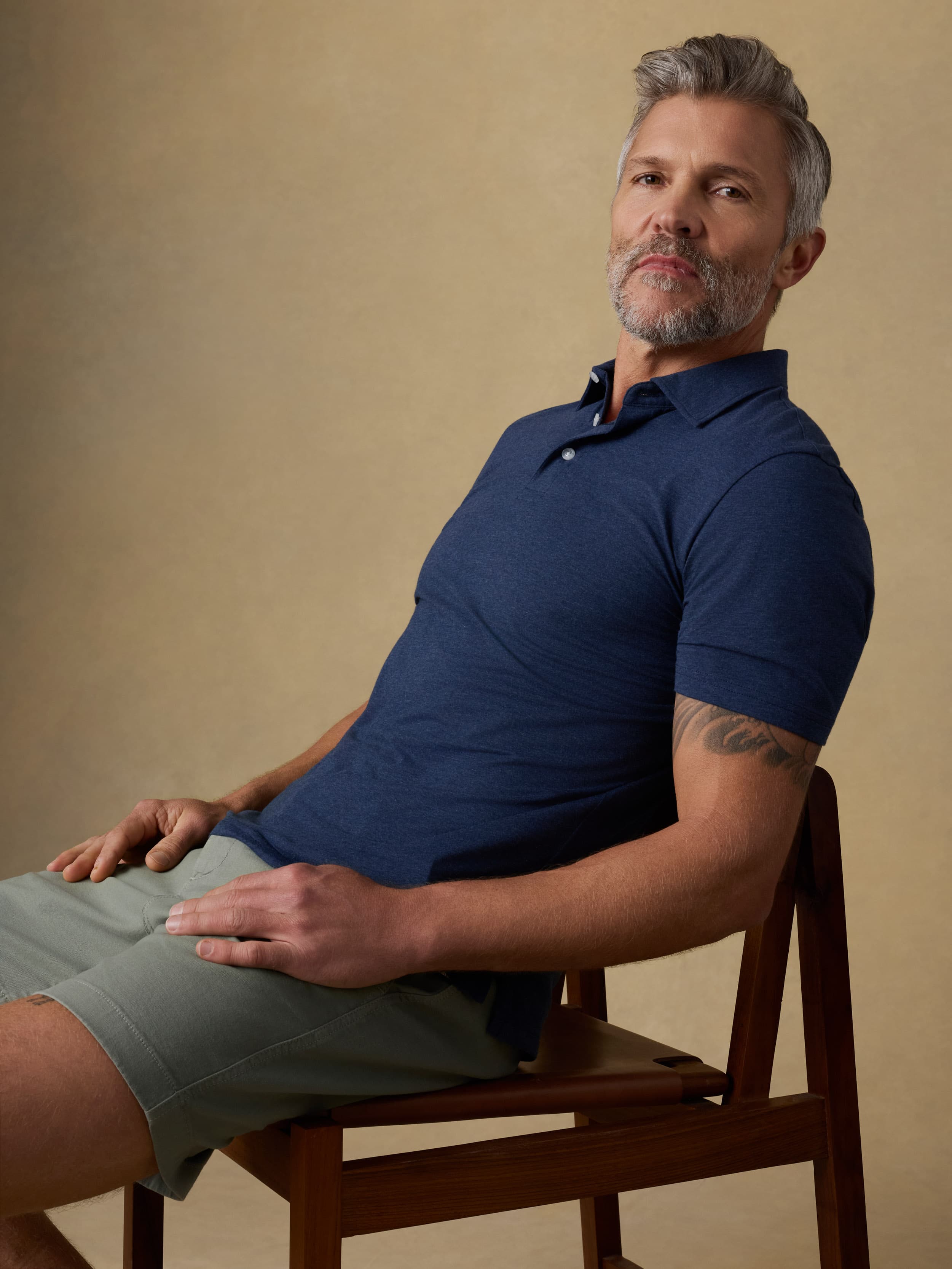 A man with gray hair and a beard sits on a wooden chair, wearing a navy blue short-sleeve polo shirt and light olive green shorts against a warm beige studio background.