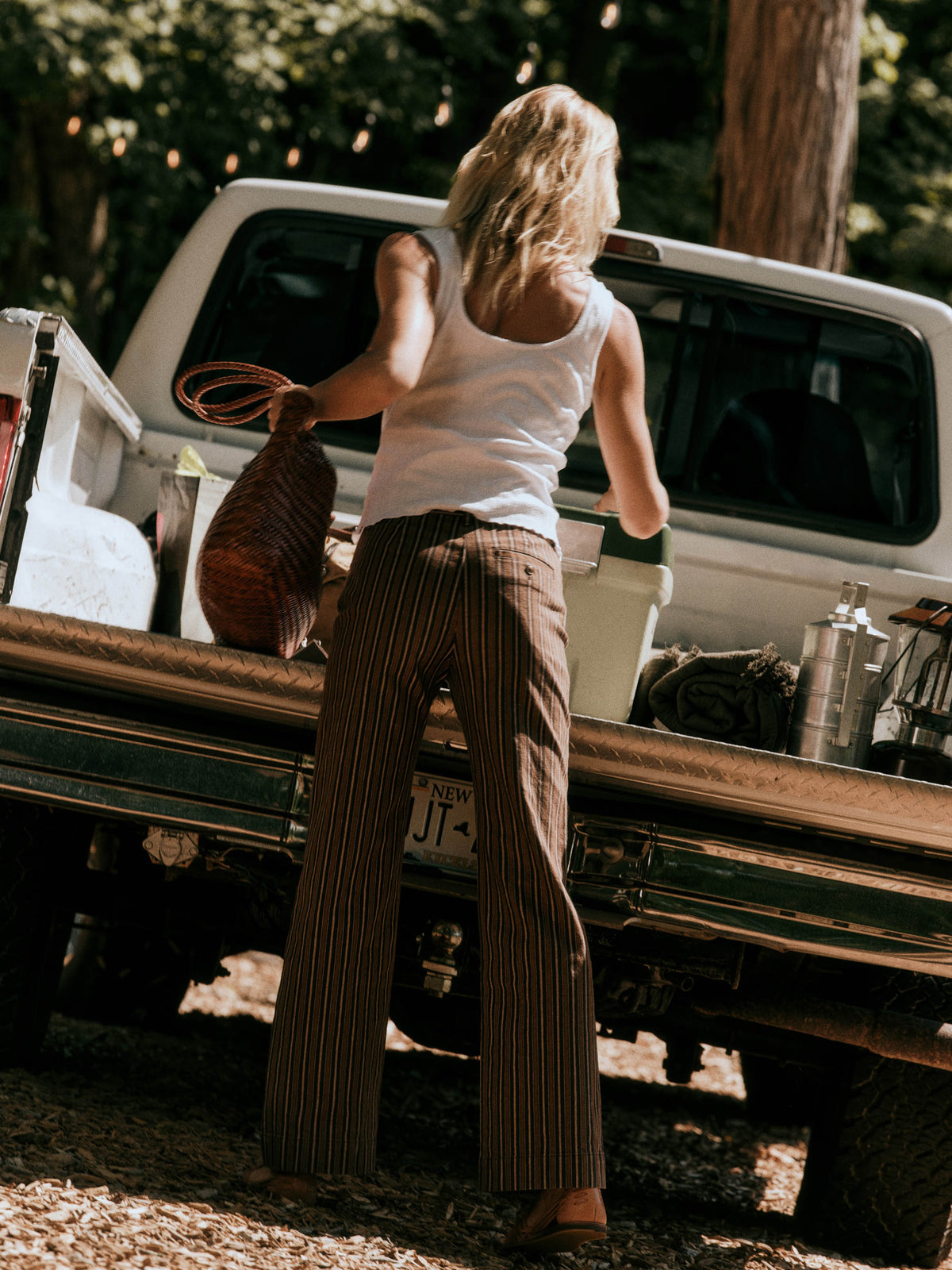A woman stands with her back to the camera, loading or unloading items from the bed of a white pickup truck in a wooded outdoor setting. She is wearing a white tank top and brown pinstriped wide-leg pants, holding a brown woven bag., image 7 of 7