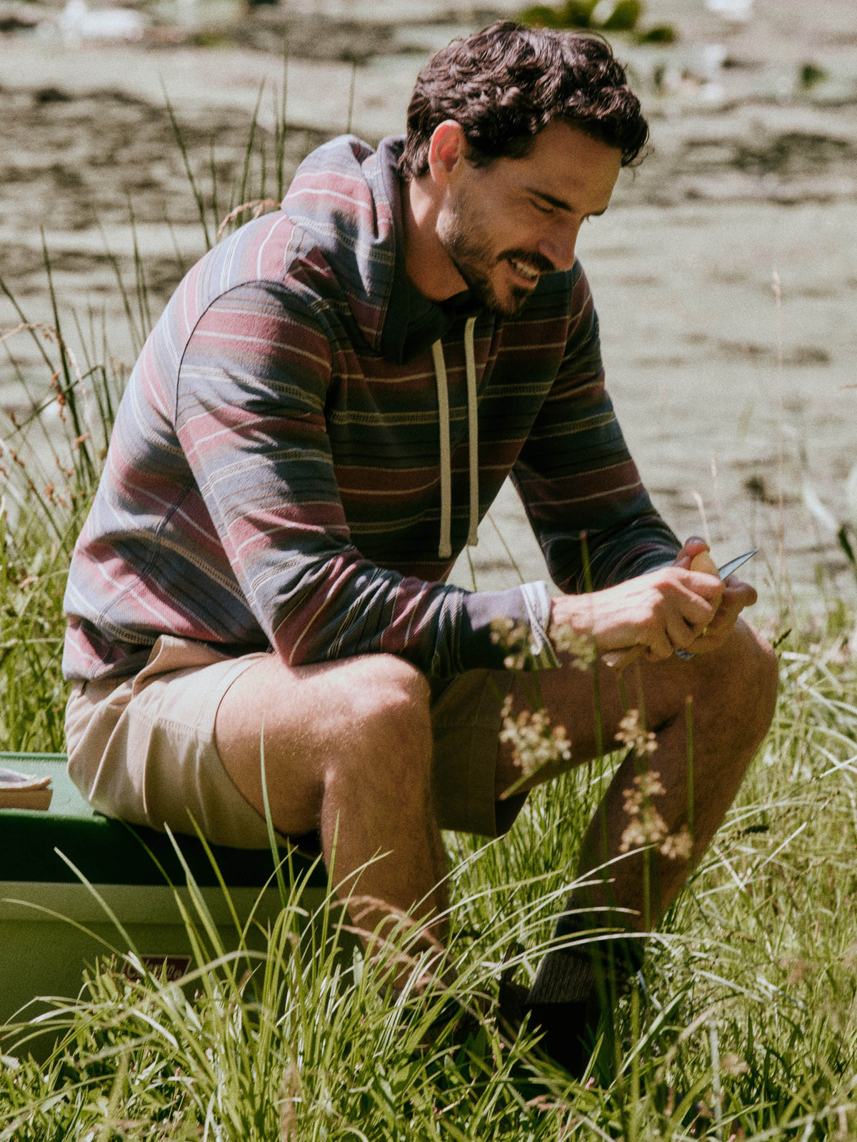 A man wearing a striped long-sleeve hoodie and beige shorts sits in tall grass outdoors, looking down at a phone., image 1 of 7