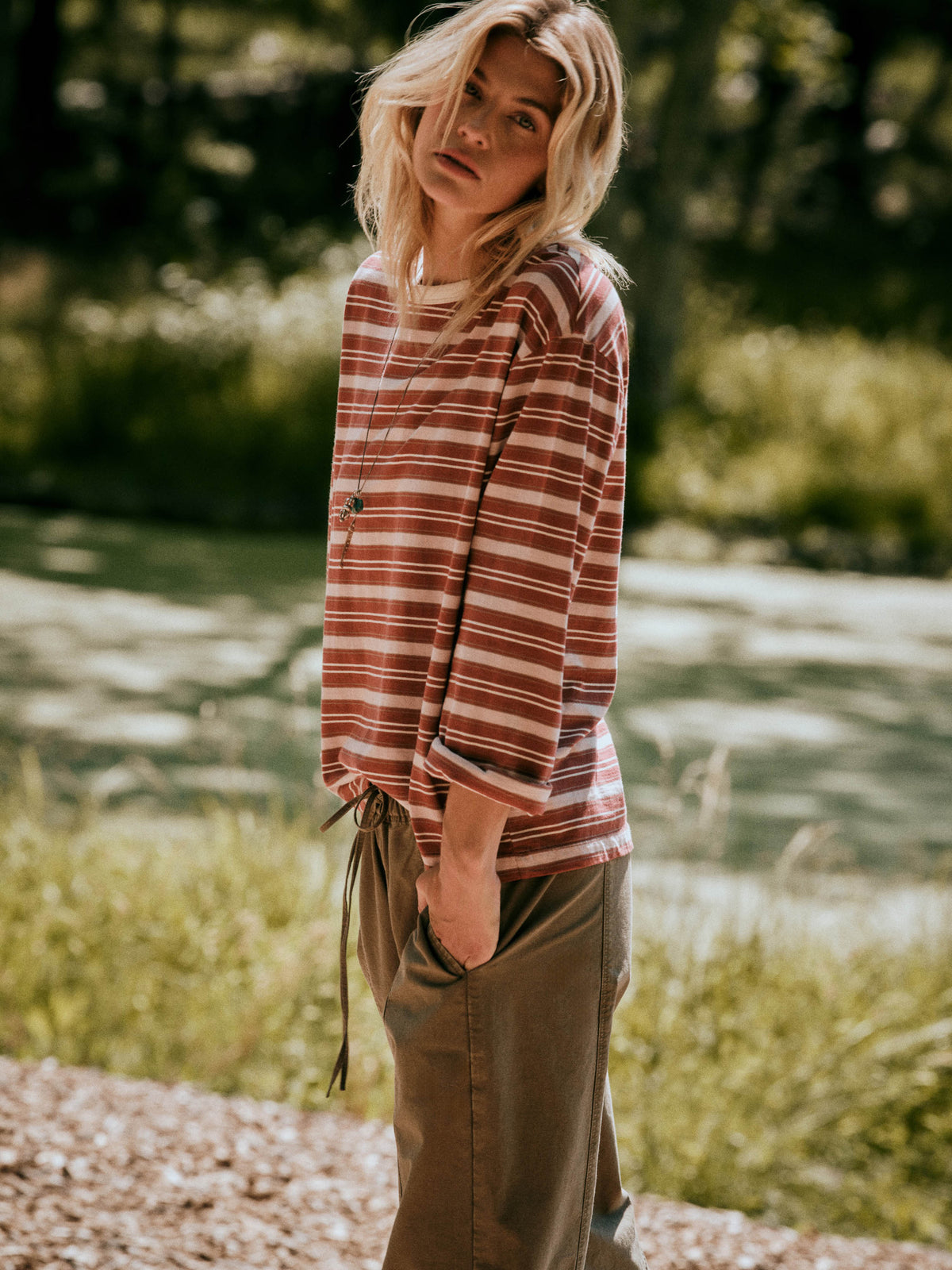A woman standing outdoors in a wooded area, wearing a red and light pink striped long-sleeve top with a drawstring hem and olive green wide-leg pants., image 1 of 7