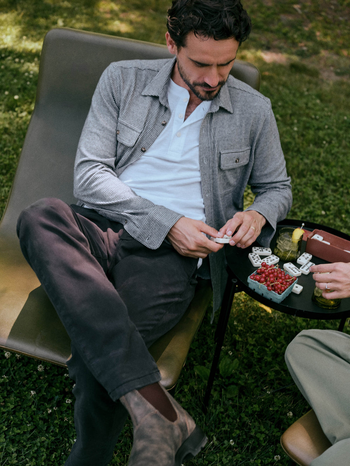 A man sits in a lounge chair outdoors on grass, wearing a light gray overshirt over a white polo shirt and dark trousers. A small table beside him holds cards and a drink., image 1 of 7