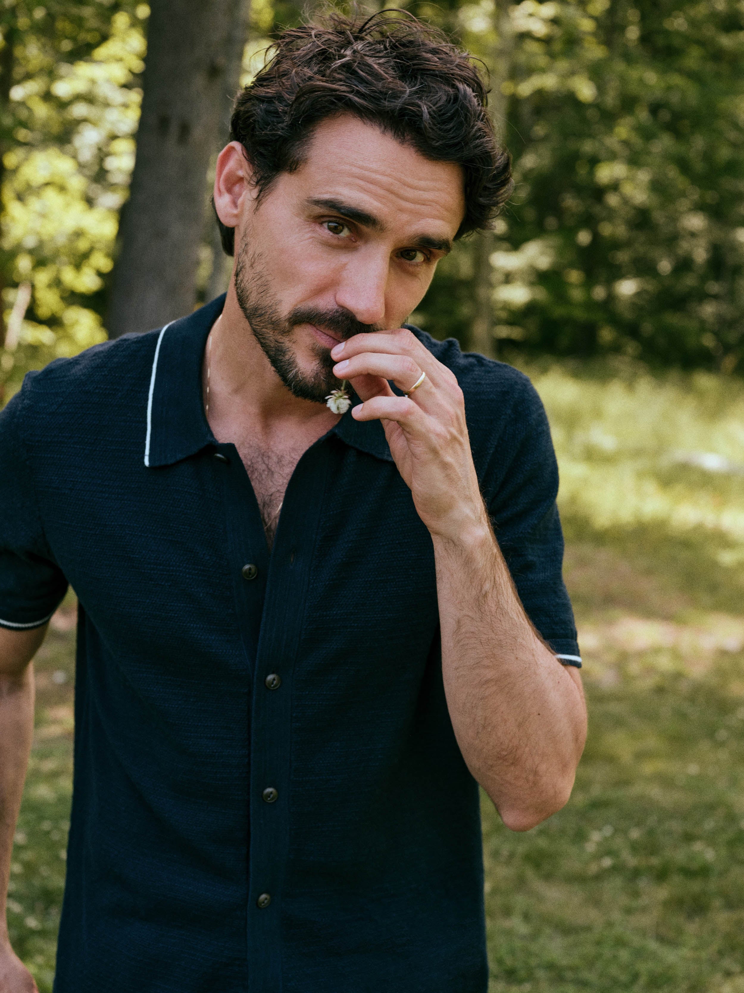 A man wearing a dark navy blue short-sleeve polo shirt with white piping on the collar and sleeve cuffs, standing outdoors in a grassy, wooded area.