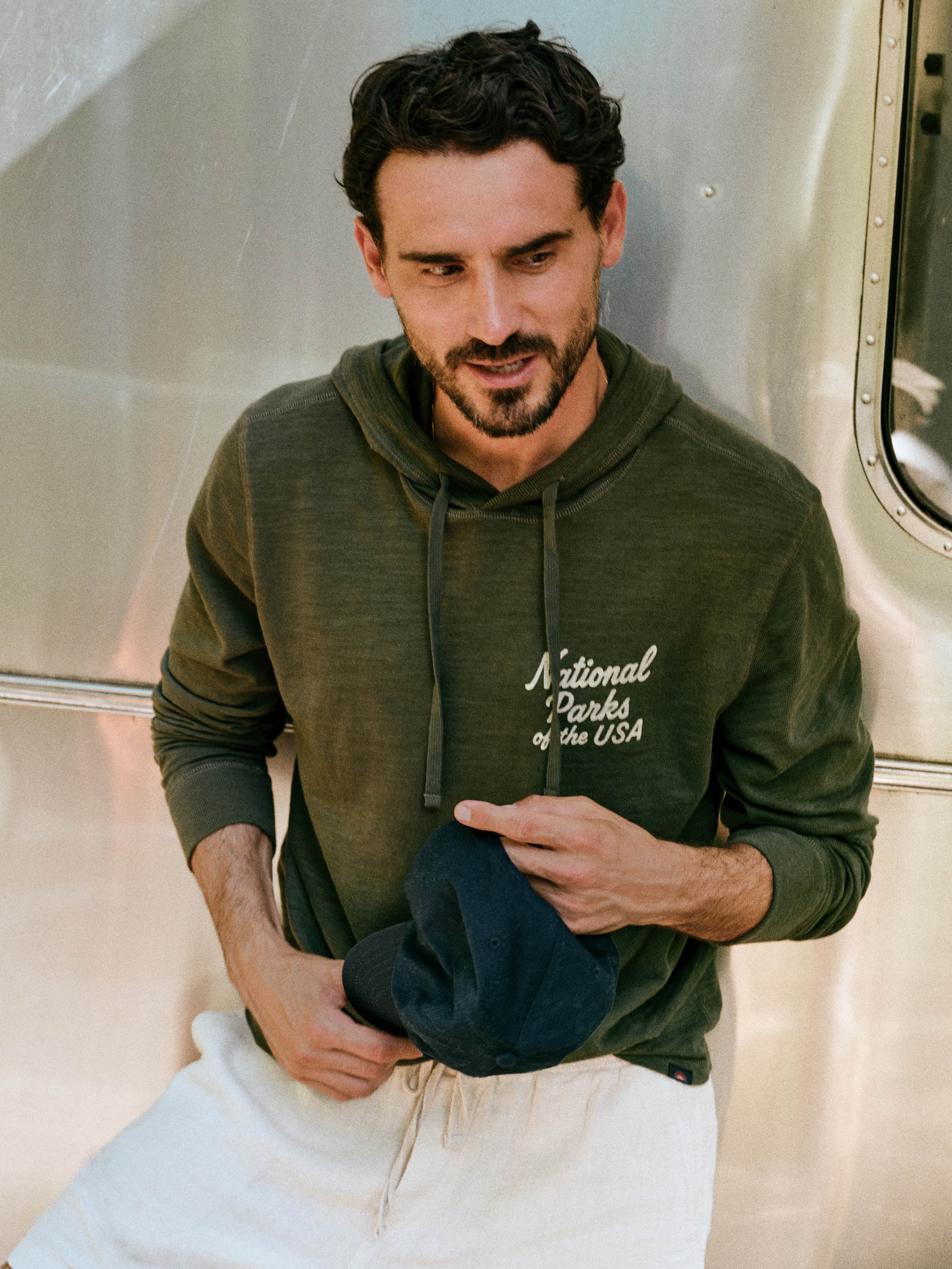 A man wearing an olive green hoodie with 'National Parks of the USA' printed in white script on the chest, paired with white sweatpants and holding a dark blue cap, posed in front of a metallic silver trailer.