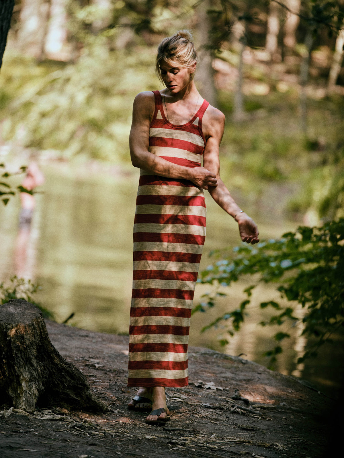 A woman wearing a red and beige horizontally striped maxi dress stands on a dirt path near a body of water in a wooded outdoor setting., image 1 of 6