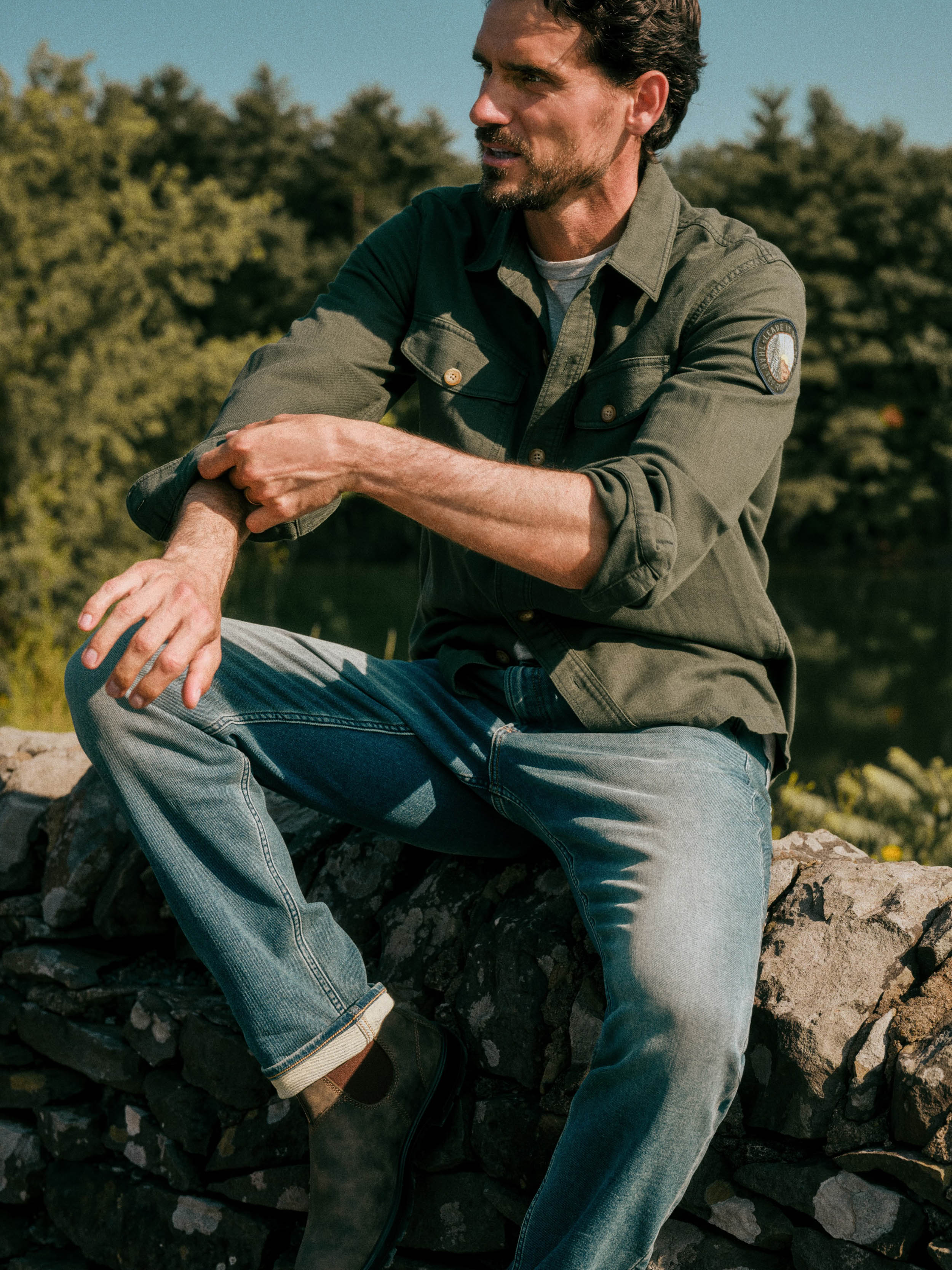 A man sitting outdoors on a stone wall wearing an olive green button-up shirt and blue jeans, with trees and blue sky in the background.