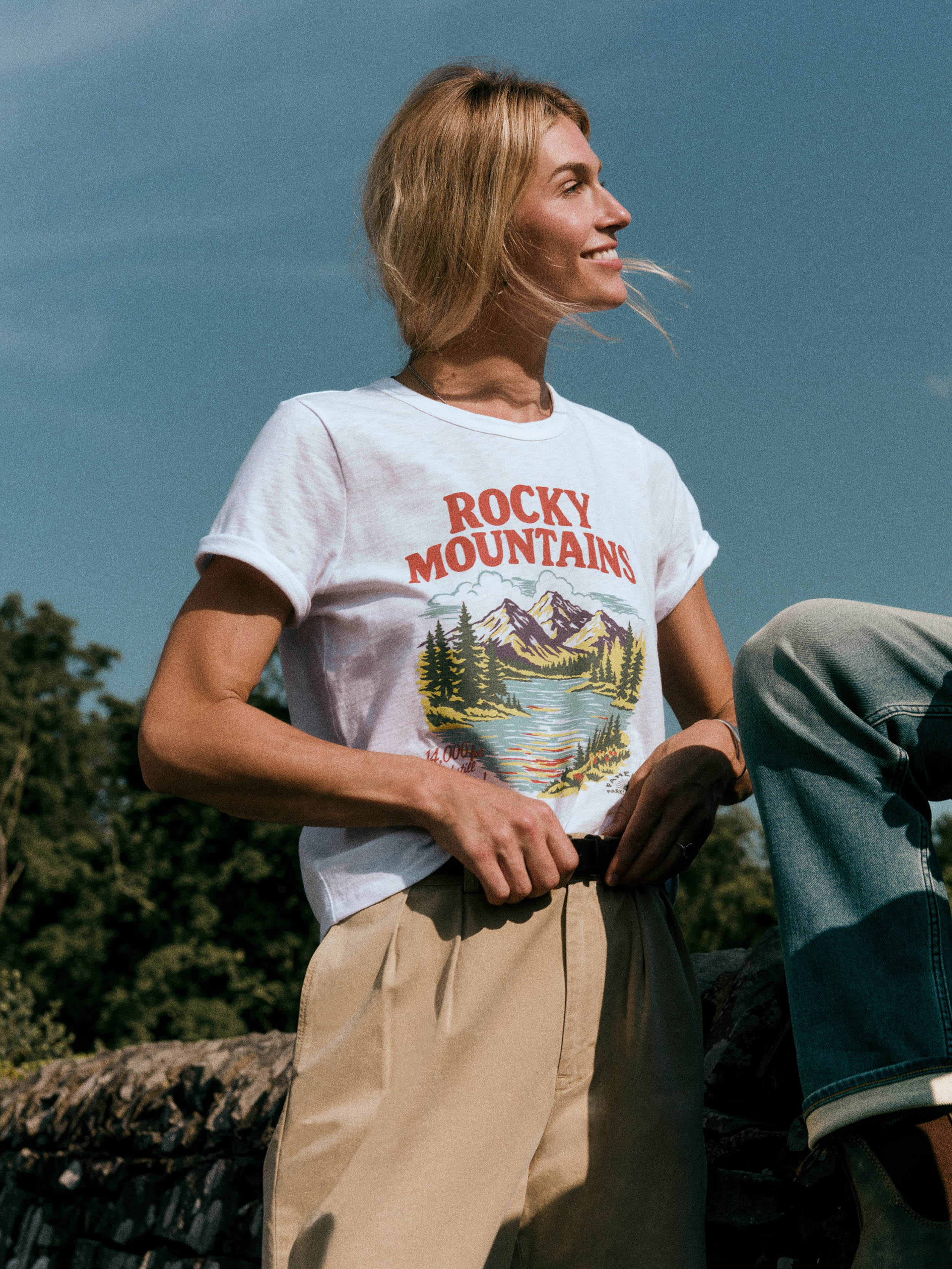 A woman wearing a white graphic t-shirt with 'ROCKY MOUNTAINS' and a mountain lake print, paired with beige pants, photographed outdoors against a blue sky.