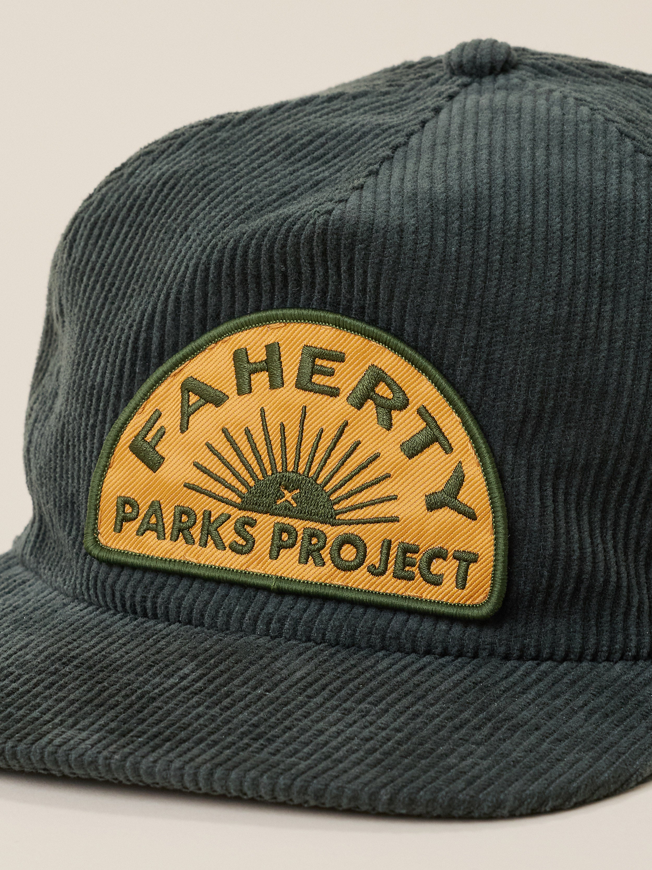 A close-up of a dark green corduroy baseball cap featuring a prominent yellow and green embroidered patch on the front that reads 'FAHERTY x PARKS PROJECT'.