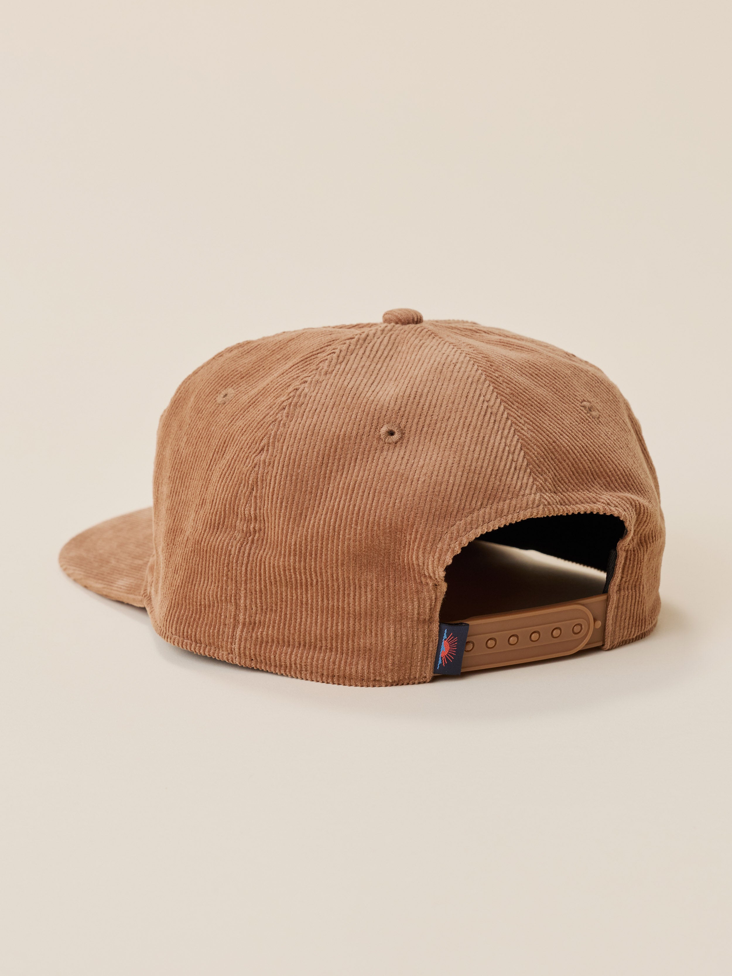 A brown corduroy snapback cap shown from the back, featuring a flat brim and an adjustable strap closure, set against a solid light beige background.