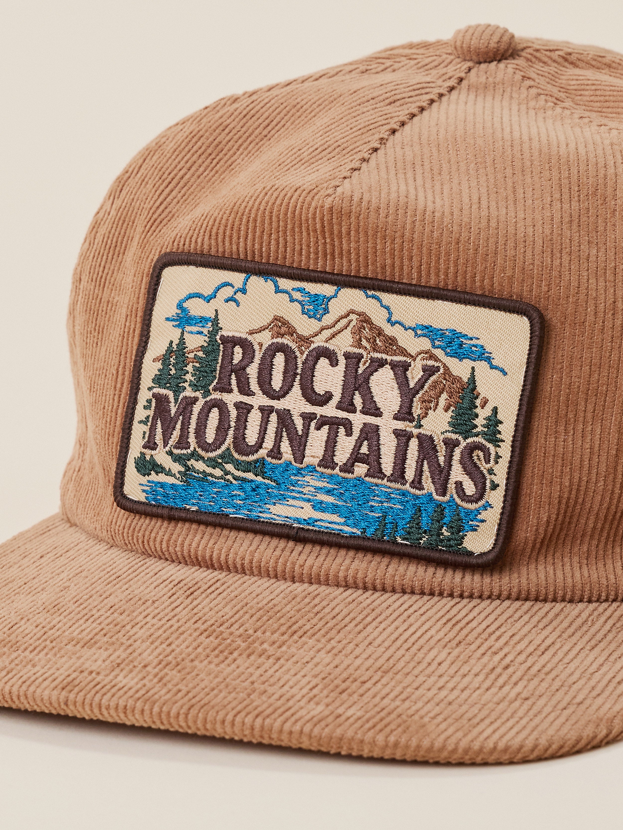 A close-up of a tan corduroy baseball cap featuring a rectangular embroidered patch on the front with a mountain landscape and the words 'ROCKY MOUNTAINS'.