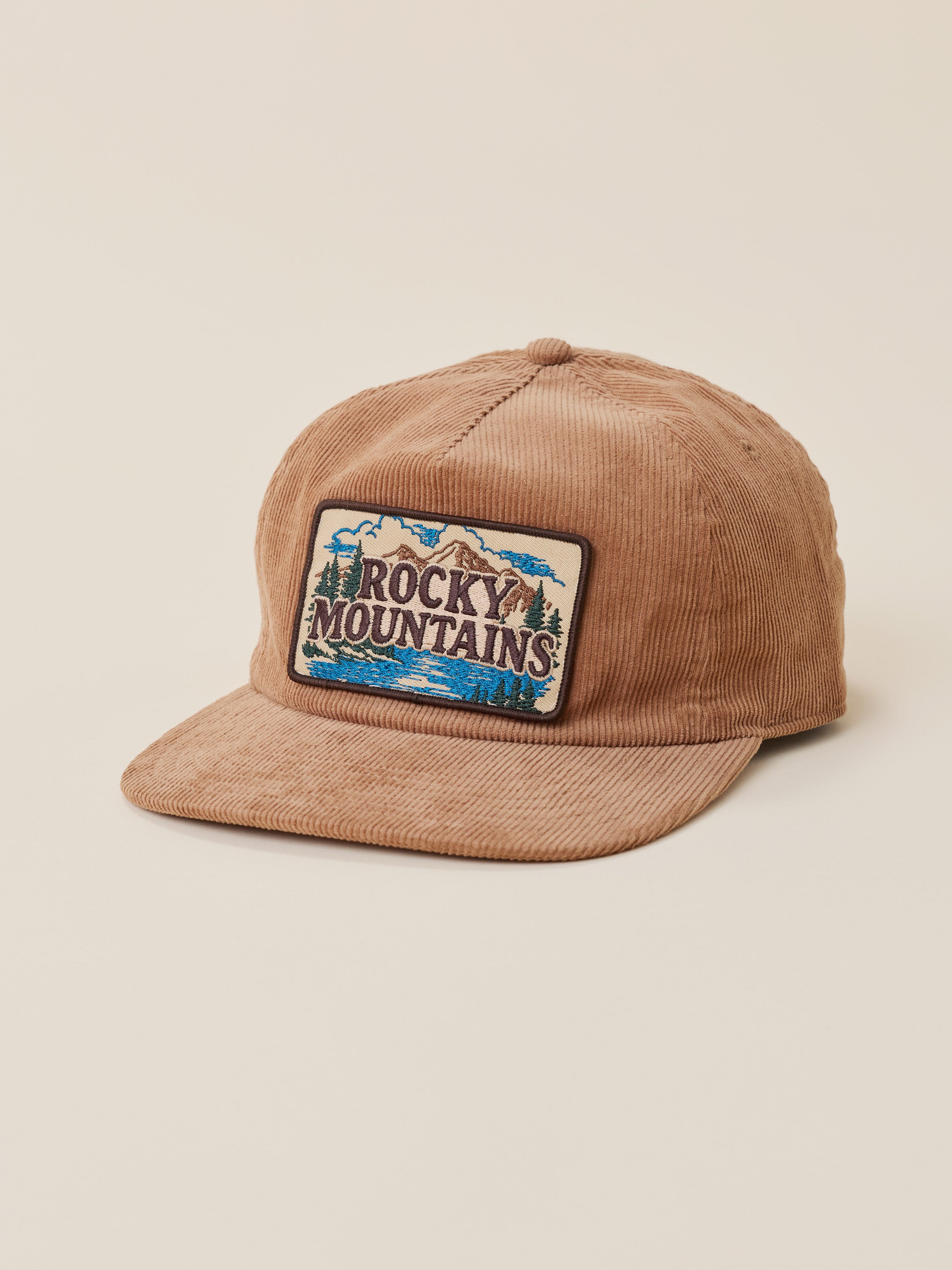 A tan corduroy baseball cap featuring a rectangular embroidered 'Rocky Mountains' patch on the front, set against a solid light beige background.