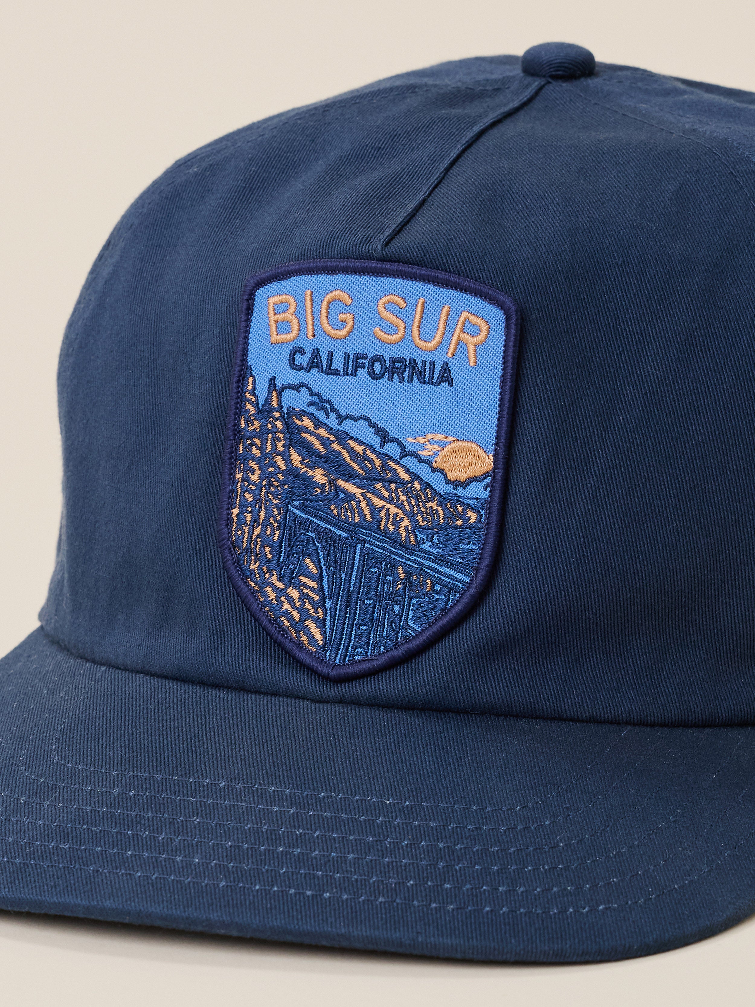 A close-up of a navy blue baseball cap featuring a shield-shaped embroidered patch on the front with the text 'BIG SUR CALIFORNIA' and a mountain landscape illustration.