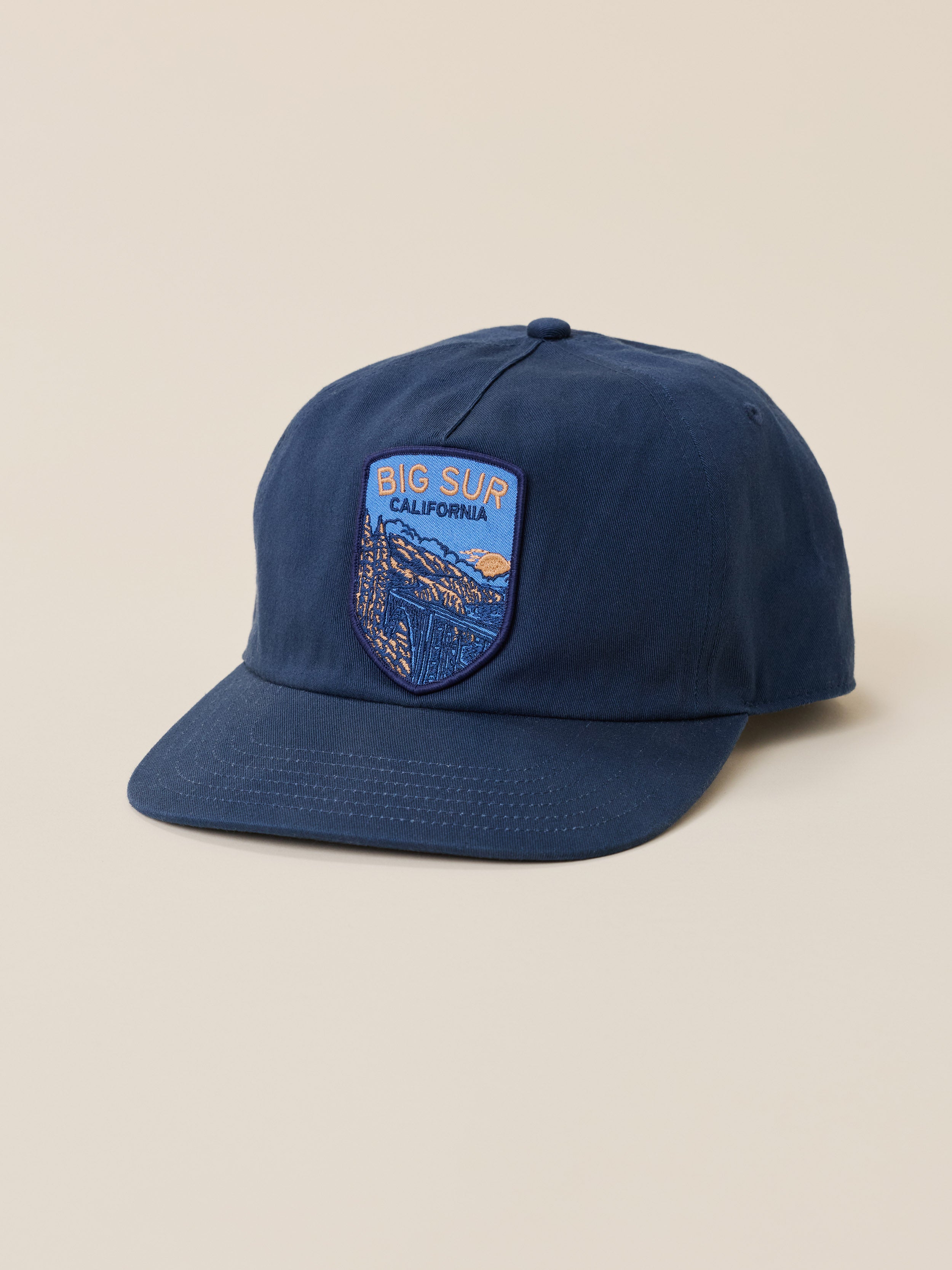A navy blue baseball cap with an embroidered shield patch on the front that reads 'BIG SUR CALIFORNIA' and depicts a scenic landscape. The cap is photographed on a solid, warm beige background.