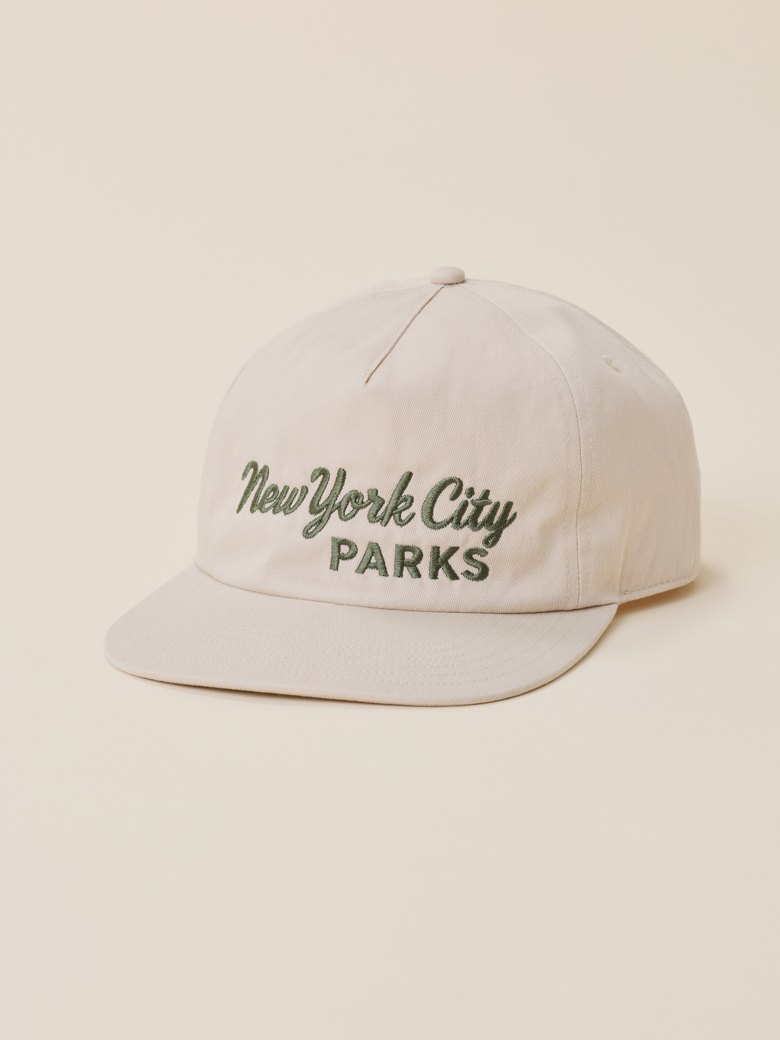 A light beige baseball cap with 'New York City Parks' embroidered on the front in dark green, set against a solid cream background.