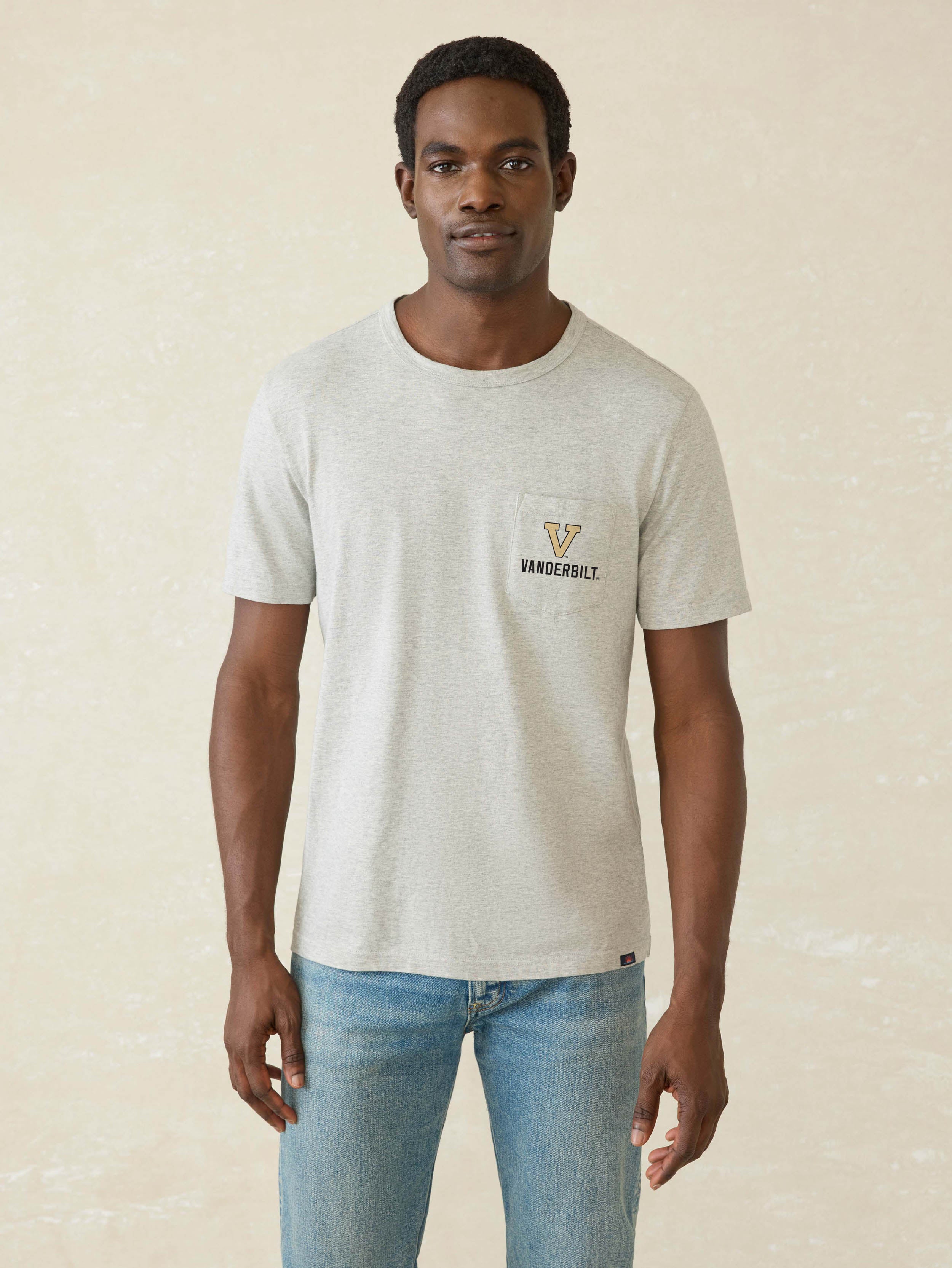 A man wearing a light heather gray t-shirt with a small gold and black 'Vanderbilt' logo on the left chest, paired with blue jeans, standing against a textured beige background.