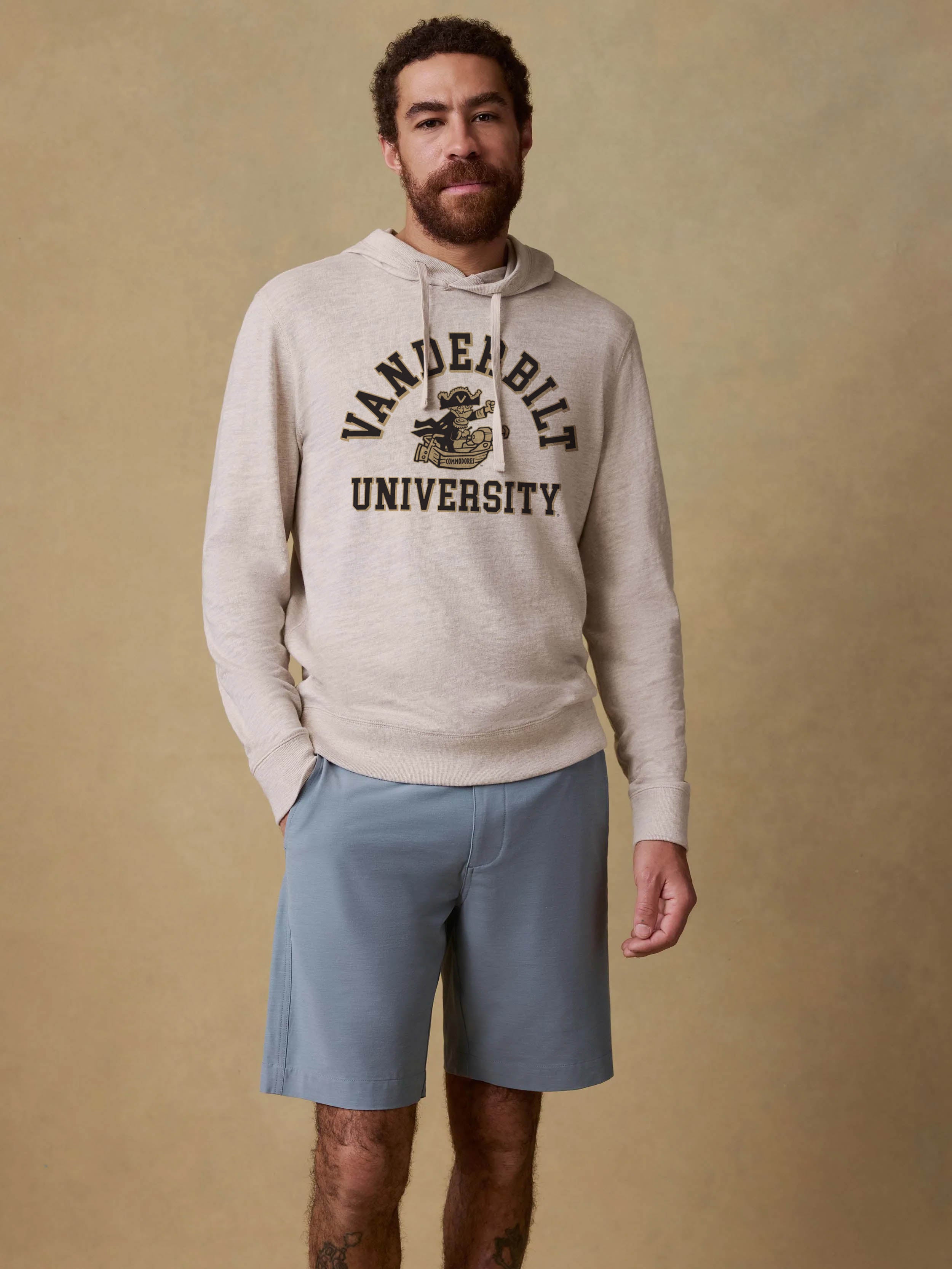A man wearing a light beige heathered hoodie with a black 'VANDERBILT UNIVERSITY' graphic on the chest, paired with light blue shorts, standing against a textured beige studio backdrop.