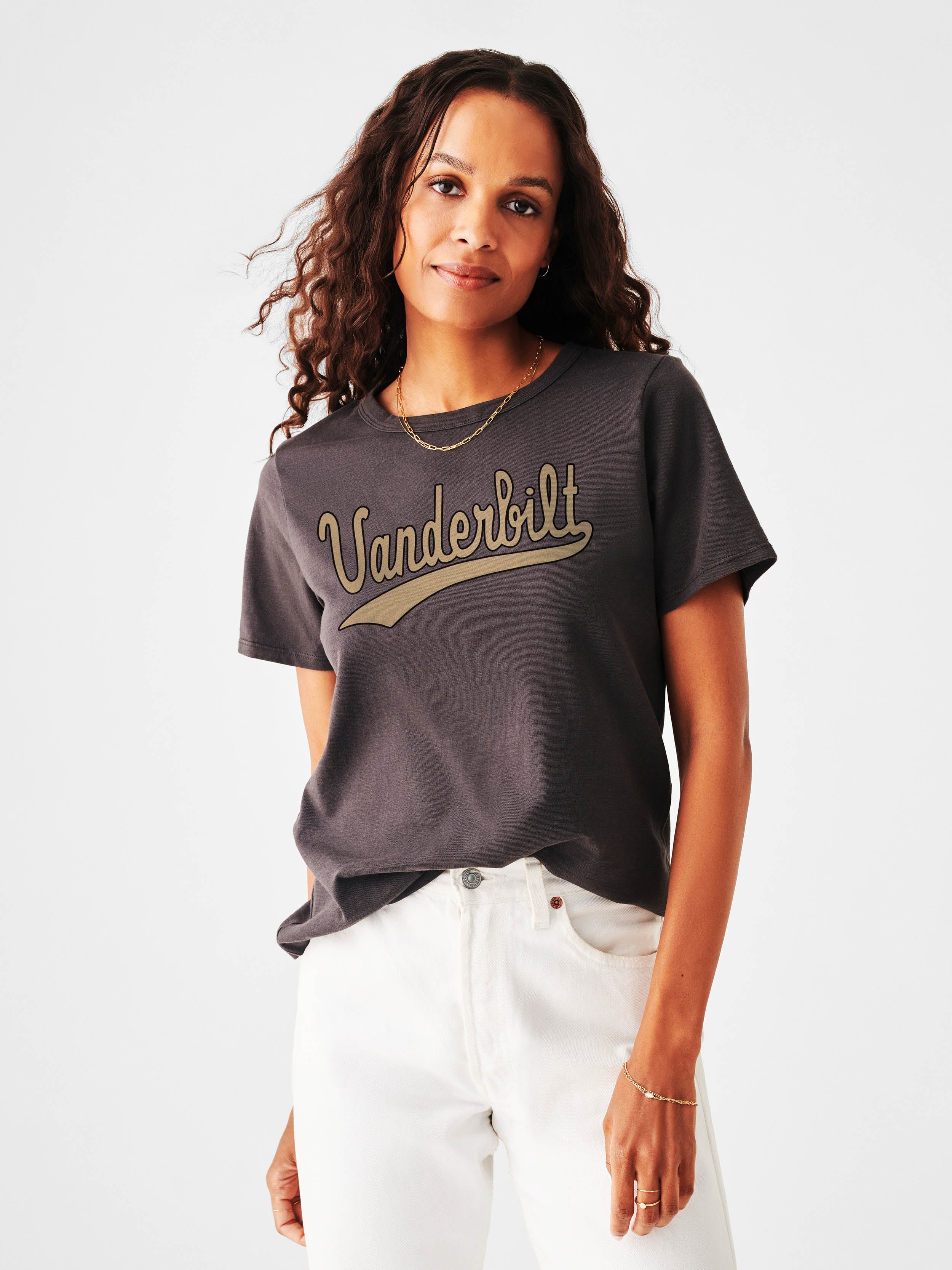 A woman wearing a dark gray t-shirt with a gold 'Vanderbilt' script graphic across the chest, paired with white pants, photographed against a light gray studio background.