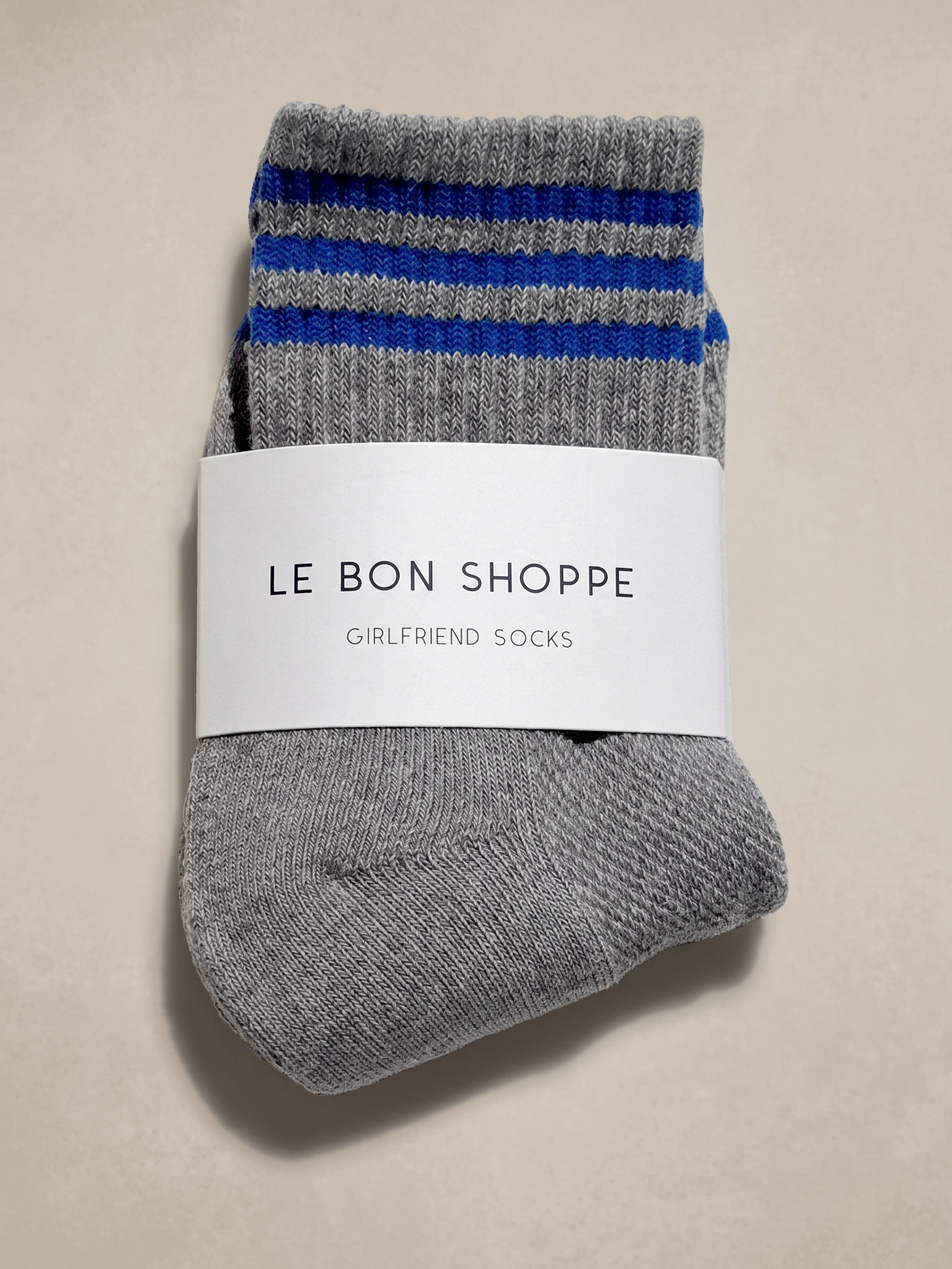 A pair of gray knit socks with blue and gray striped cuffs, wrapped in a white paper band that reads 'Le Bon Shoppe Girlfriend Socks', on a beige background.