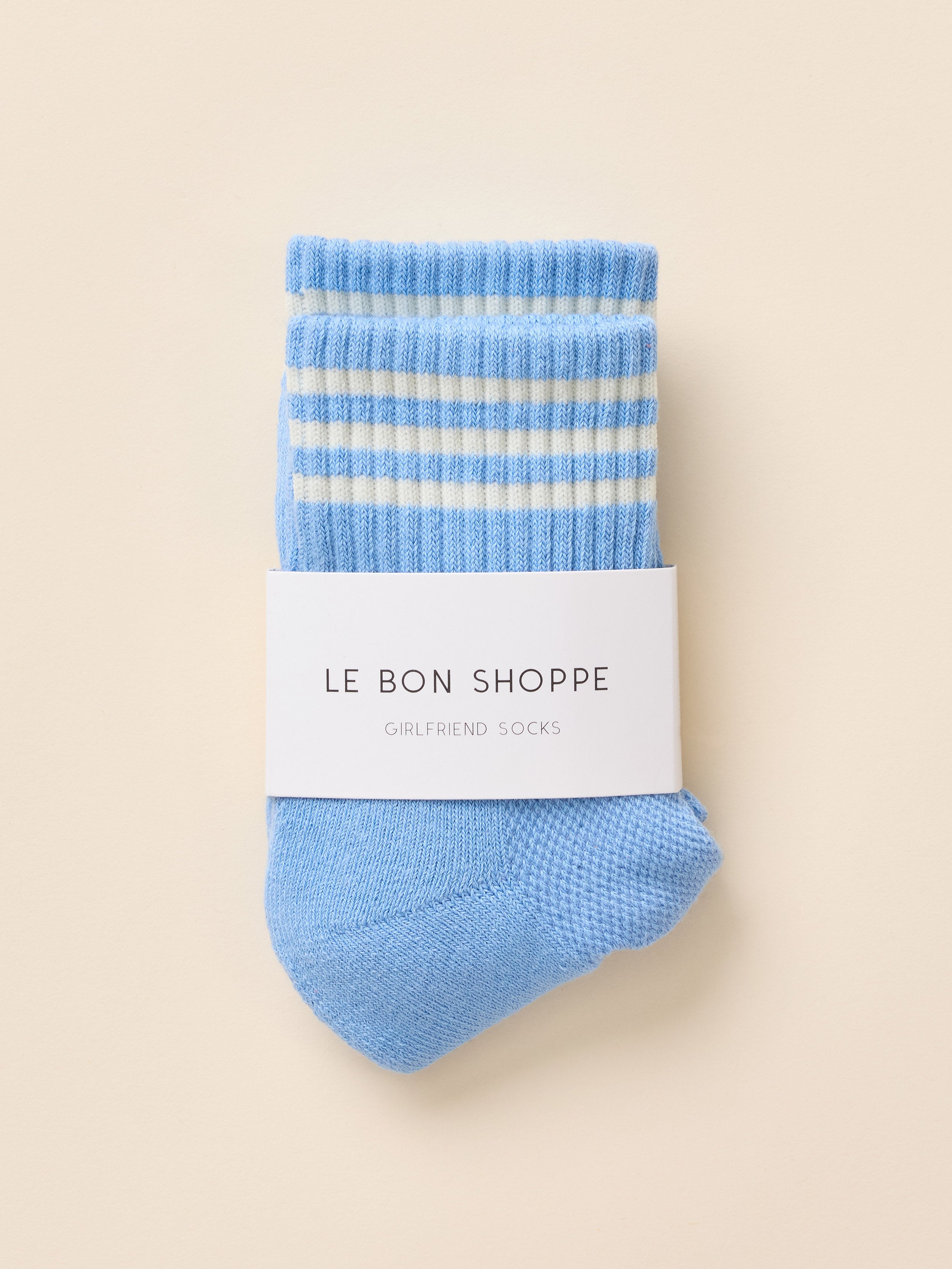 A single light blue knit sock with white horizontal stripes near the cuff, wrapped in a white paper band that reads 'LE BON SHOPPE' and 'GIRLFRIEND SOCKS', on a warm beige background.