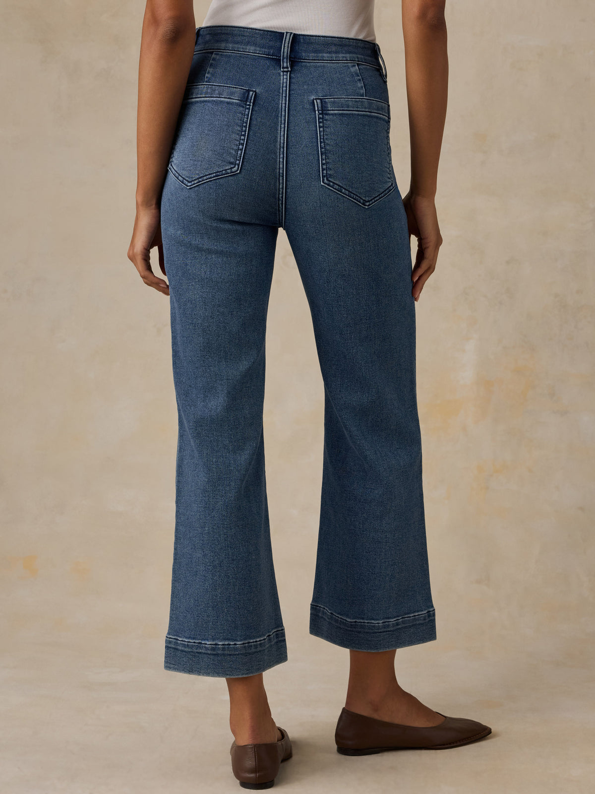 A person shown from the back wearing blue cropped flare jeans with a straight-to-wide leg, cuffed hems, and classic back pocket stitching, paired with brown flats against a textured beige studio backdrop., image 4 of 6