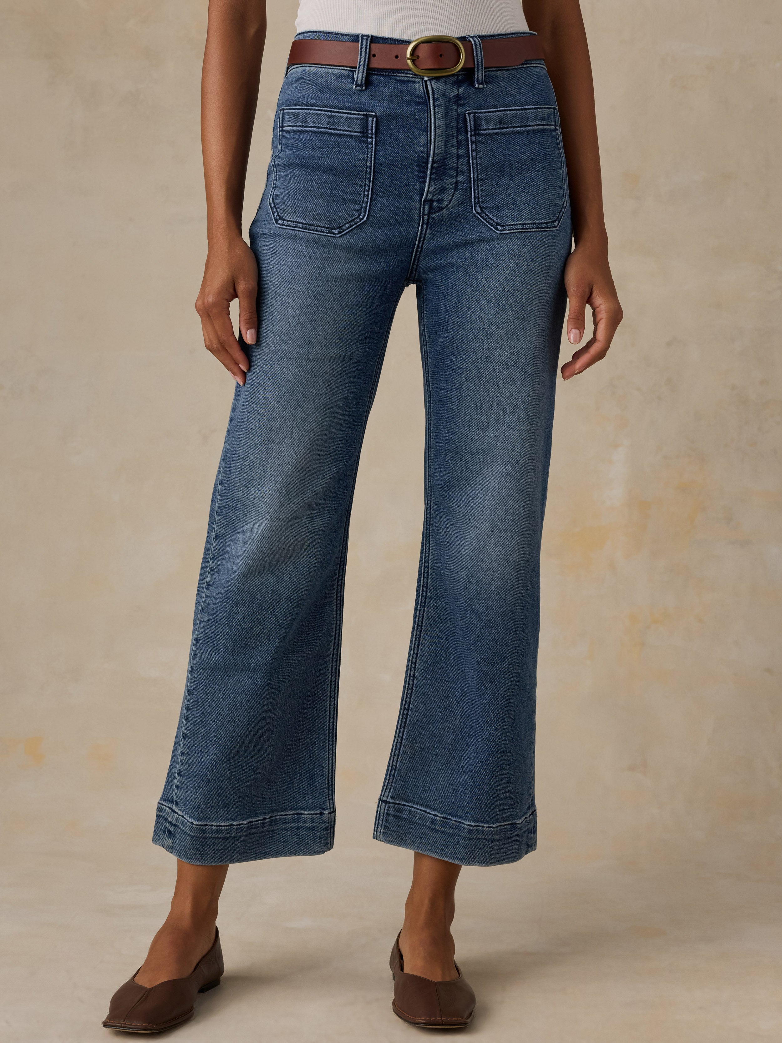 A model wearing a pair of medium-wash blue denim cropped flare jeans with a brown leather belt, styled with brown flat shoes against a textured beige studio backdrop.
