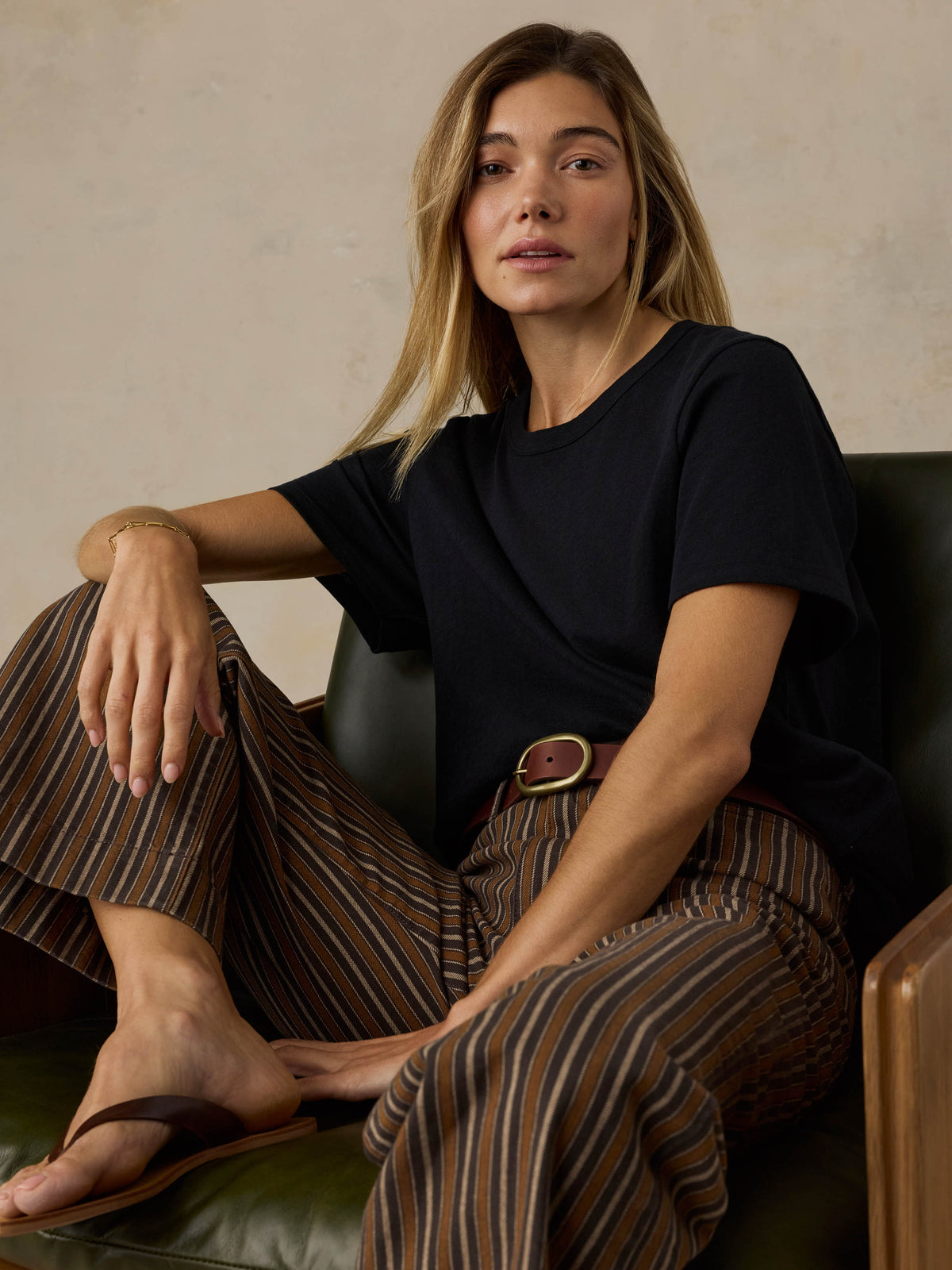Woman sitting on a wooden chair wearing a black t-shirt and brown striped pants on a beige background, image 4 of 7